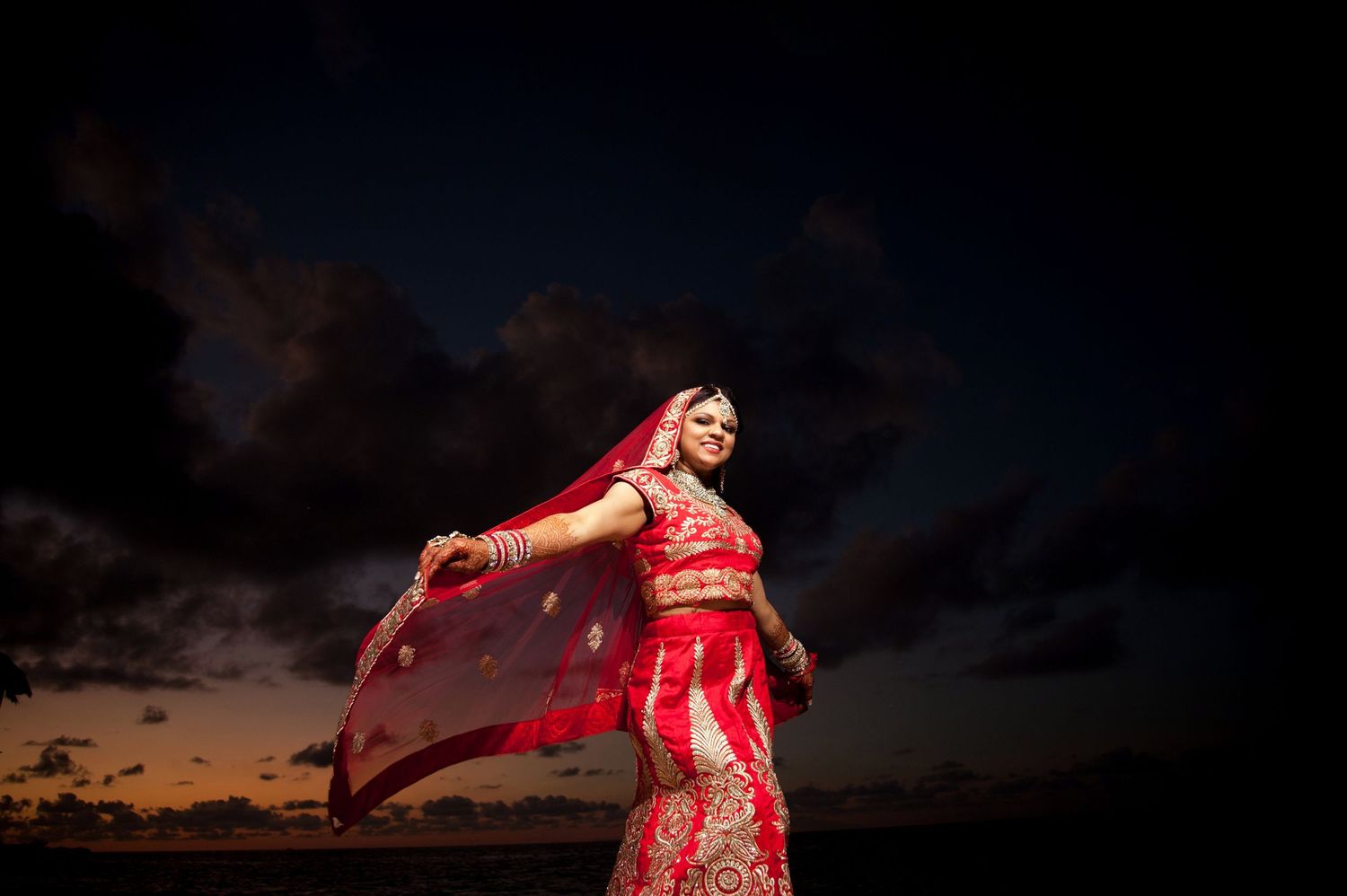 Traditional Indian bride in red sari twirls against dramatic sunset sky.