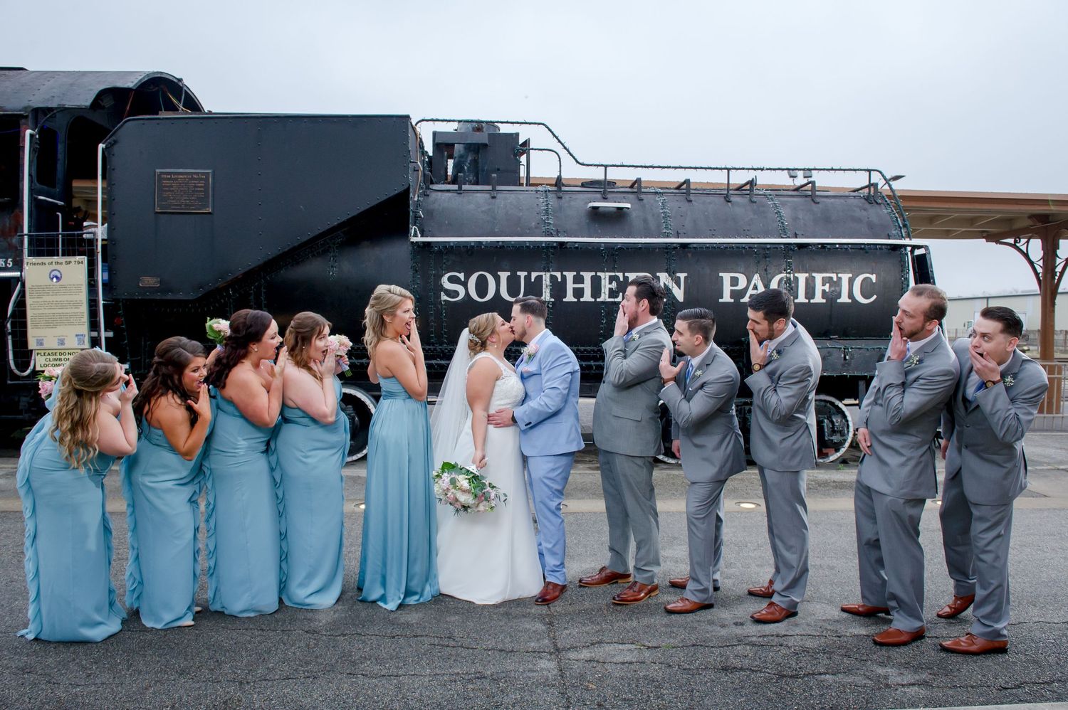 Full wedding party poses in coordinated blue and gray attire by historic train.