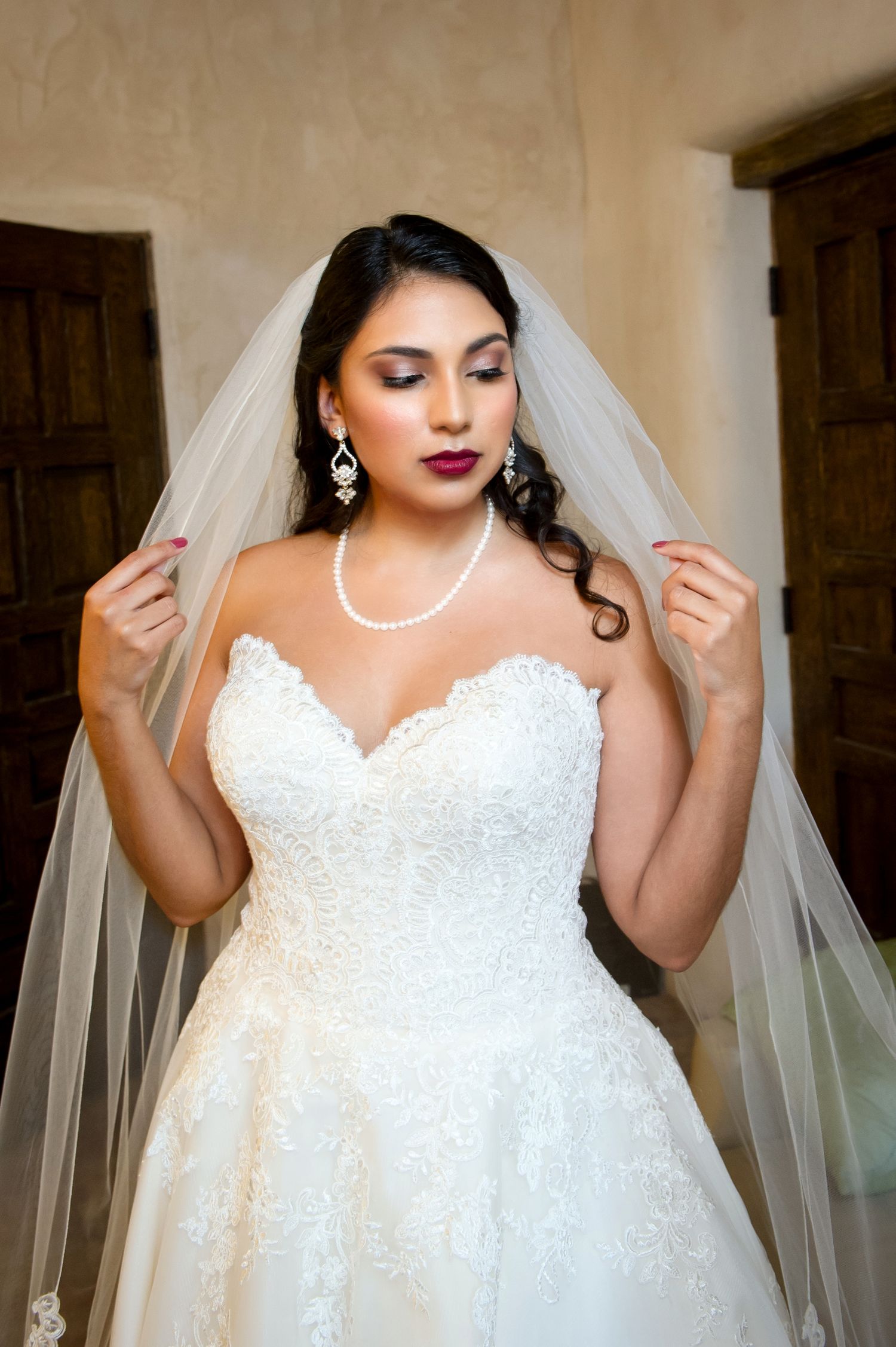 Full length portrait of bride in lace strapless gown with flowing veil against neutral wall.