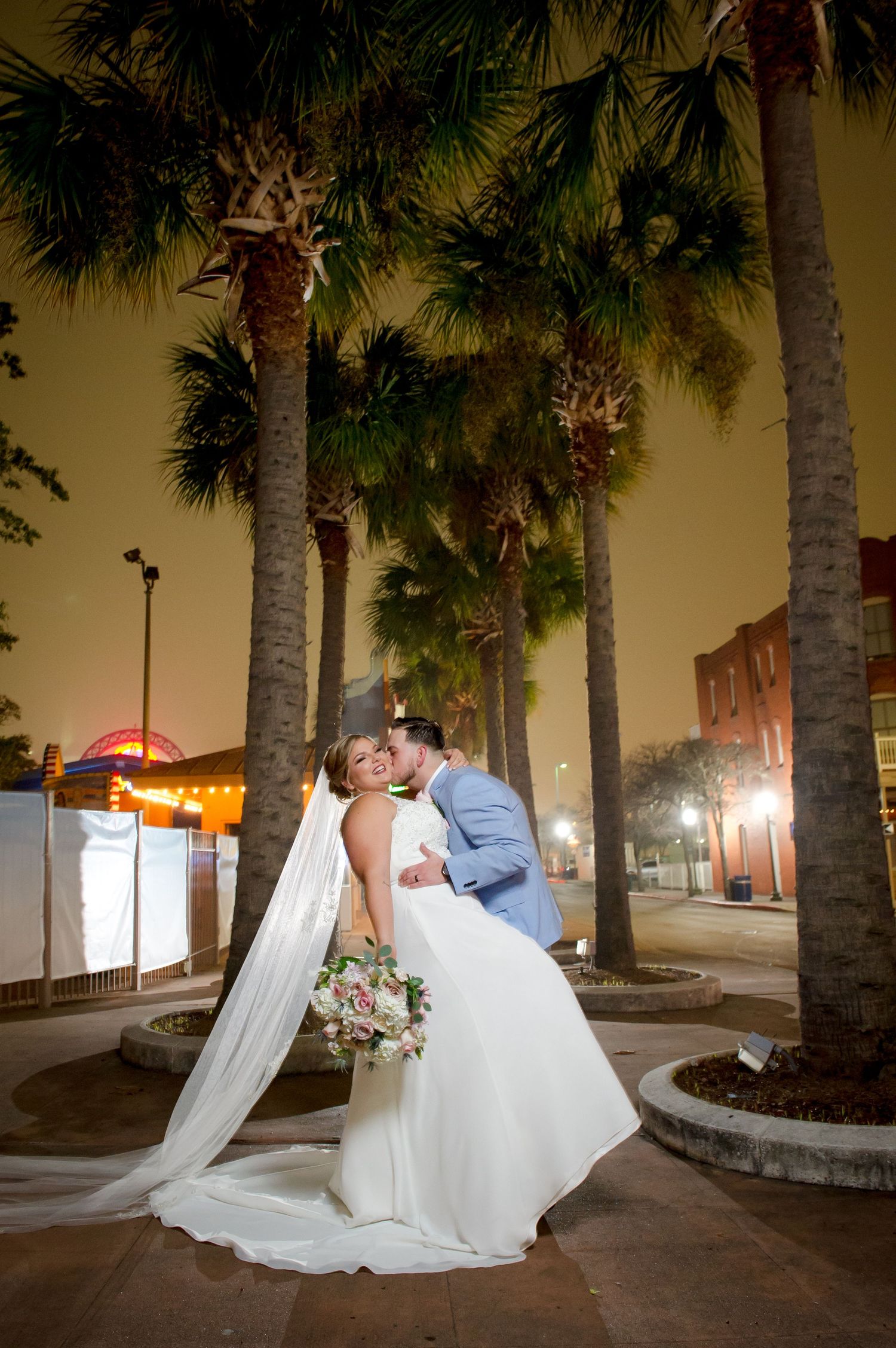 Elegant wedding couple poses under palm trees at night with city lights in background.