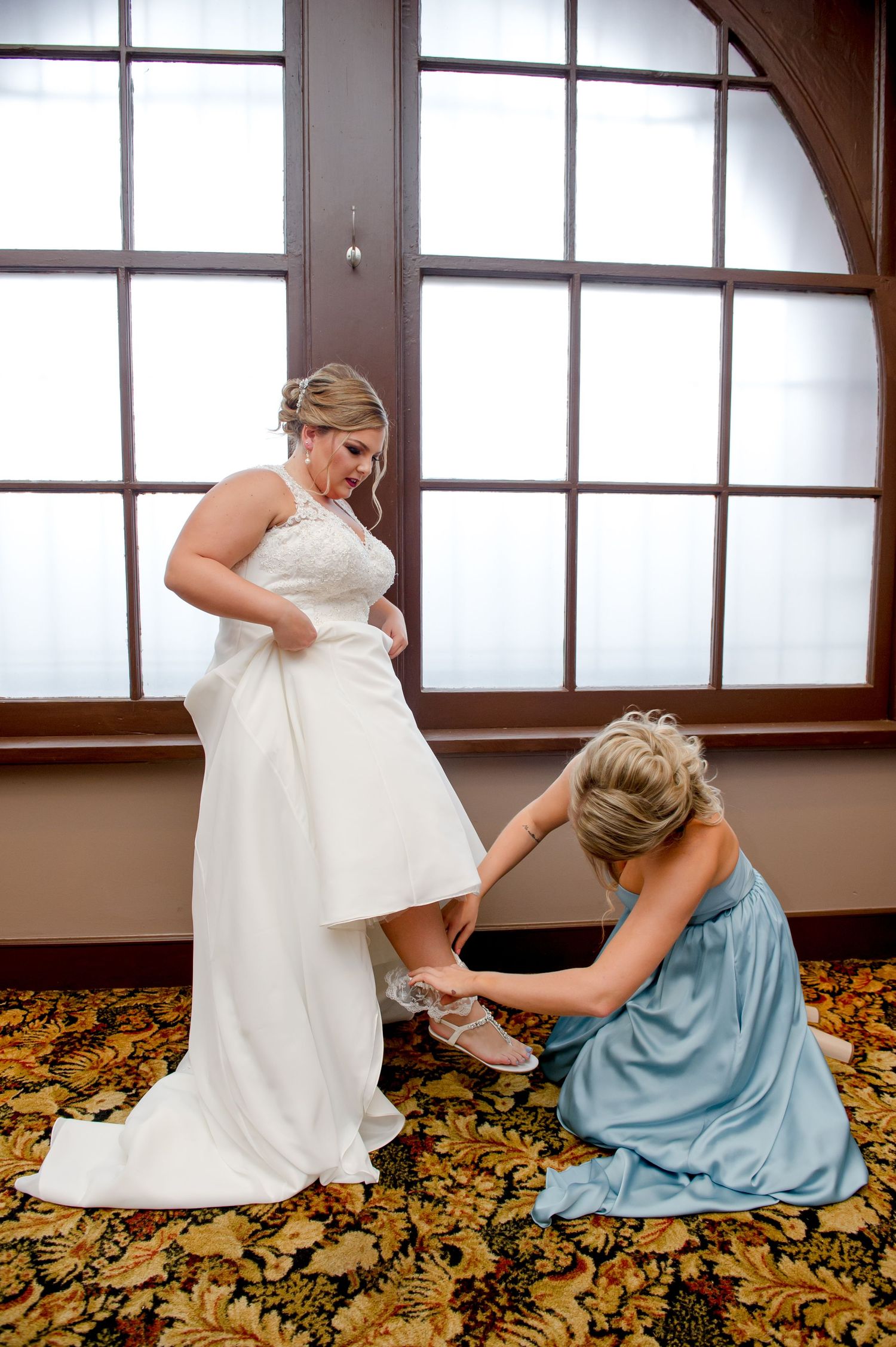 A bridesmaid helps adjust the bride's shoes on a patterned carpet.