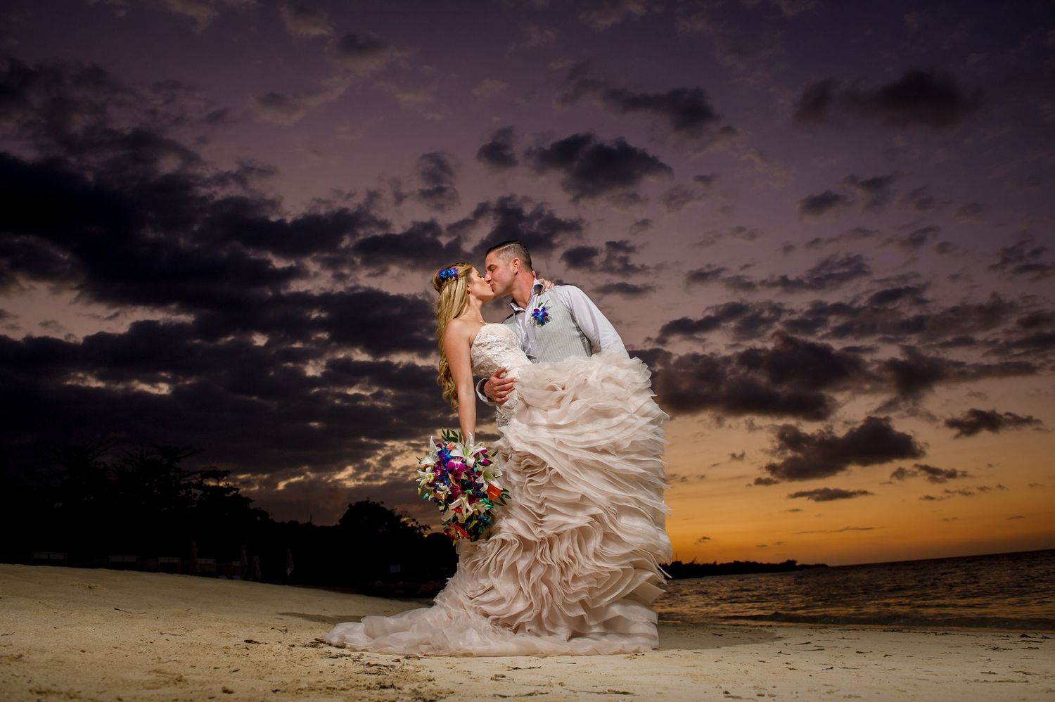Romantic beach wedding photo with dramatic sunset sky and flowing dress.