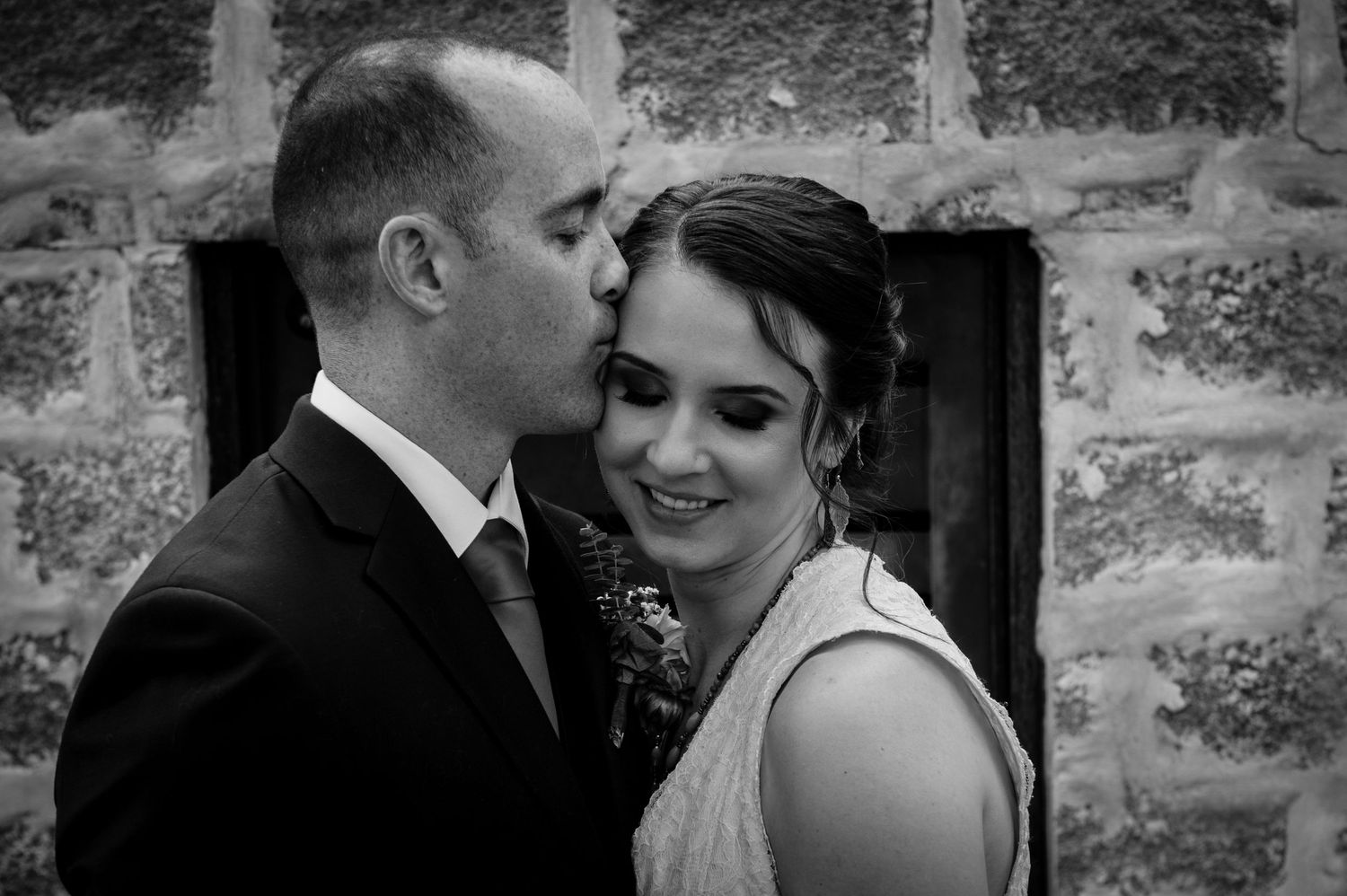 Black and white wedding photograph showing a romantic moment between newlyweds against a stone wall backdrop.