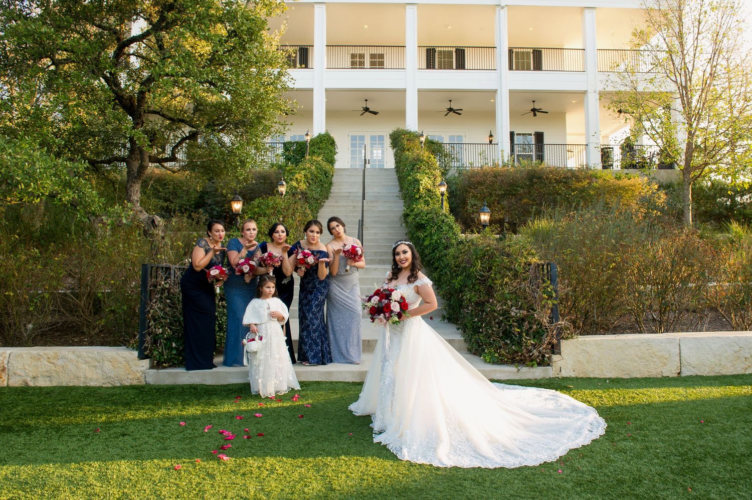 Wedding party poses in front of elegant white columned building surrounded by green landscaping.