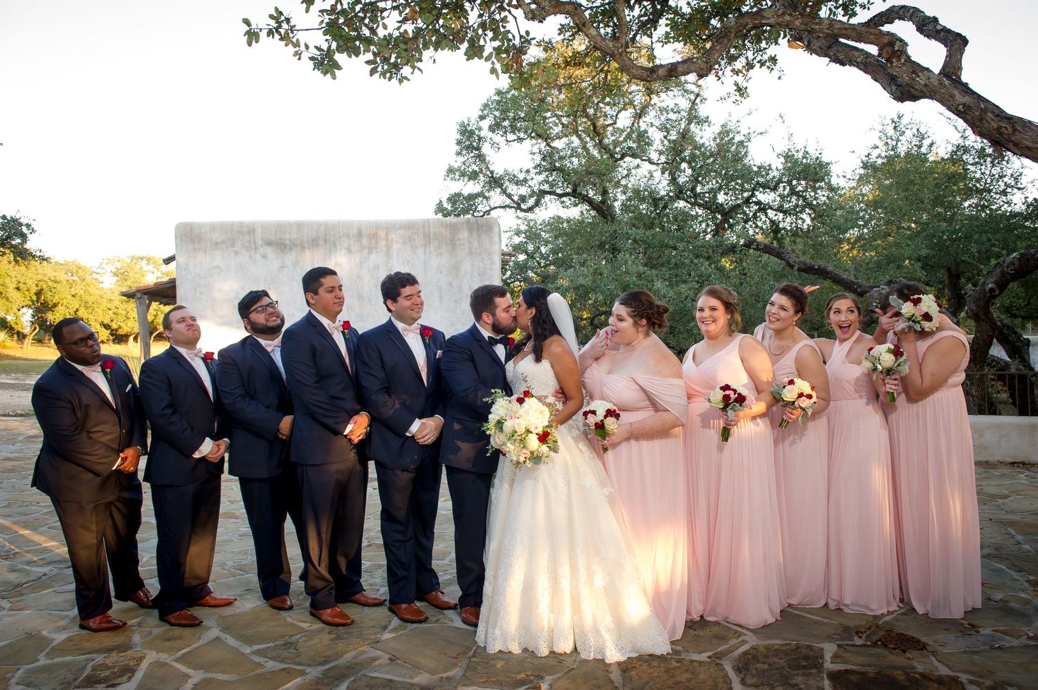 Bridal party in pink dresses and navy suits gathered for outdoor photos under oak trees.