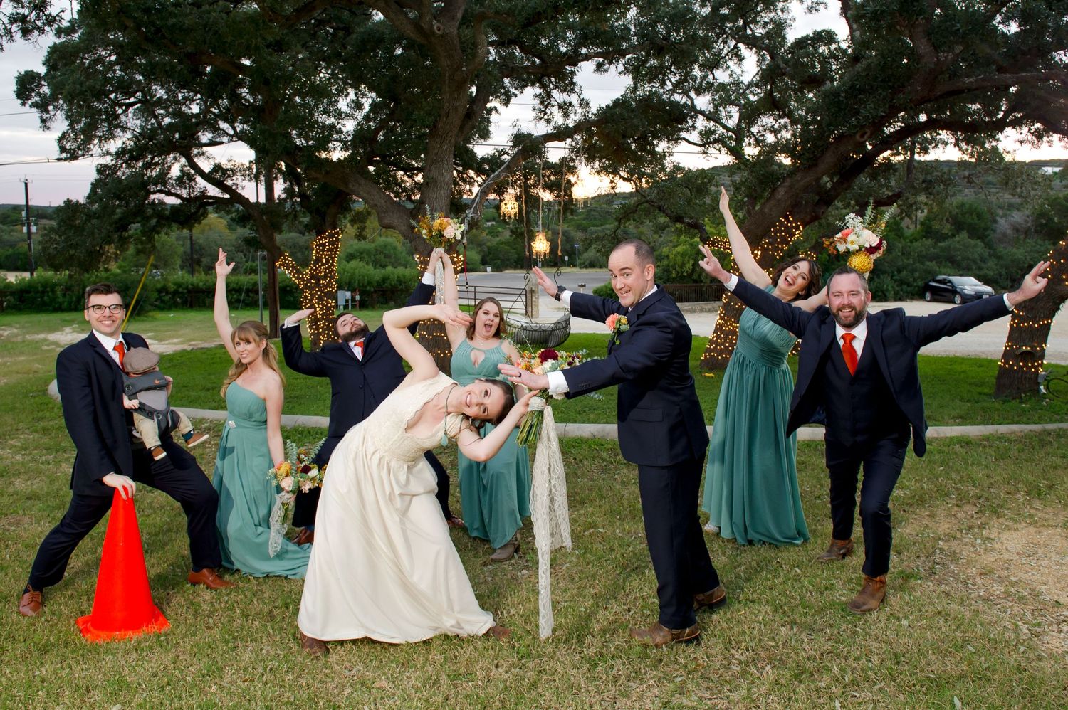 Wedding party members strike fun poses on grass under trees decorated with string lights.