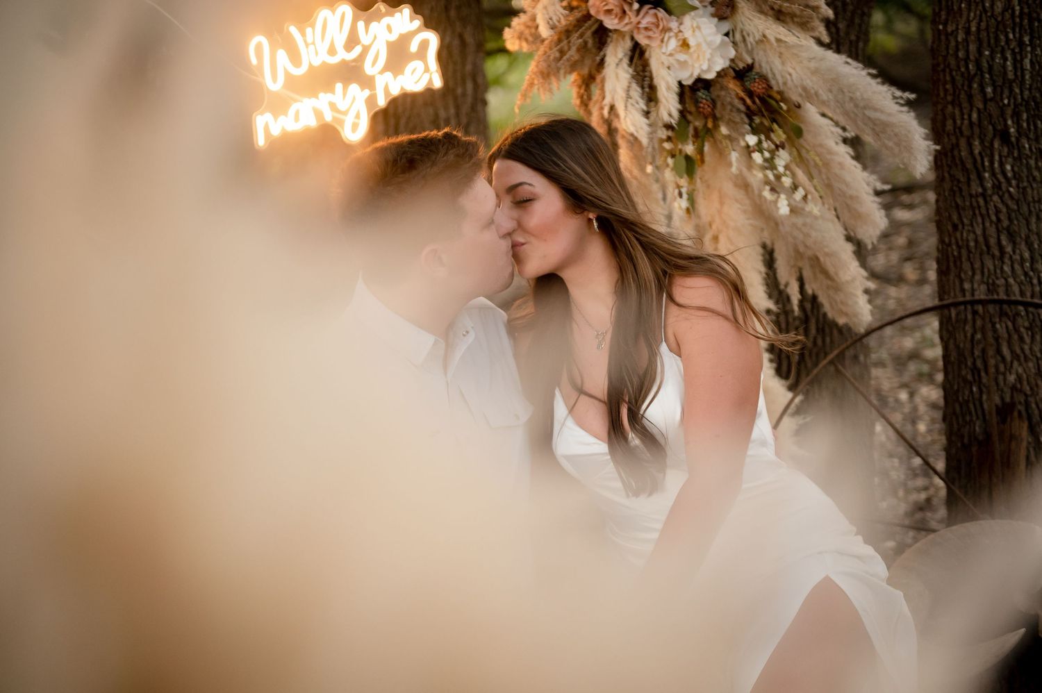 Romantic moment captured under pampas grass decorations with glowing neon sign in background.