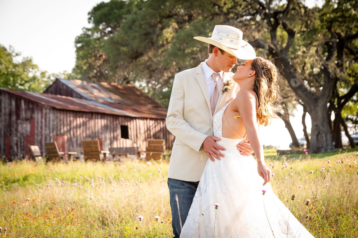 A couple in western wedding attire embraces in a rustic field with an old barn in the background.