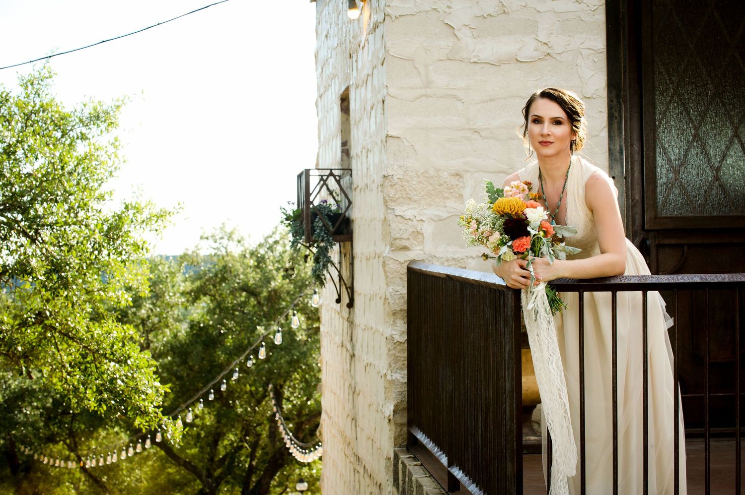 Bride in a white wedding dress holds a sunflower bouquet while standing on a rustic stone balcony at sunset.