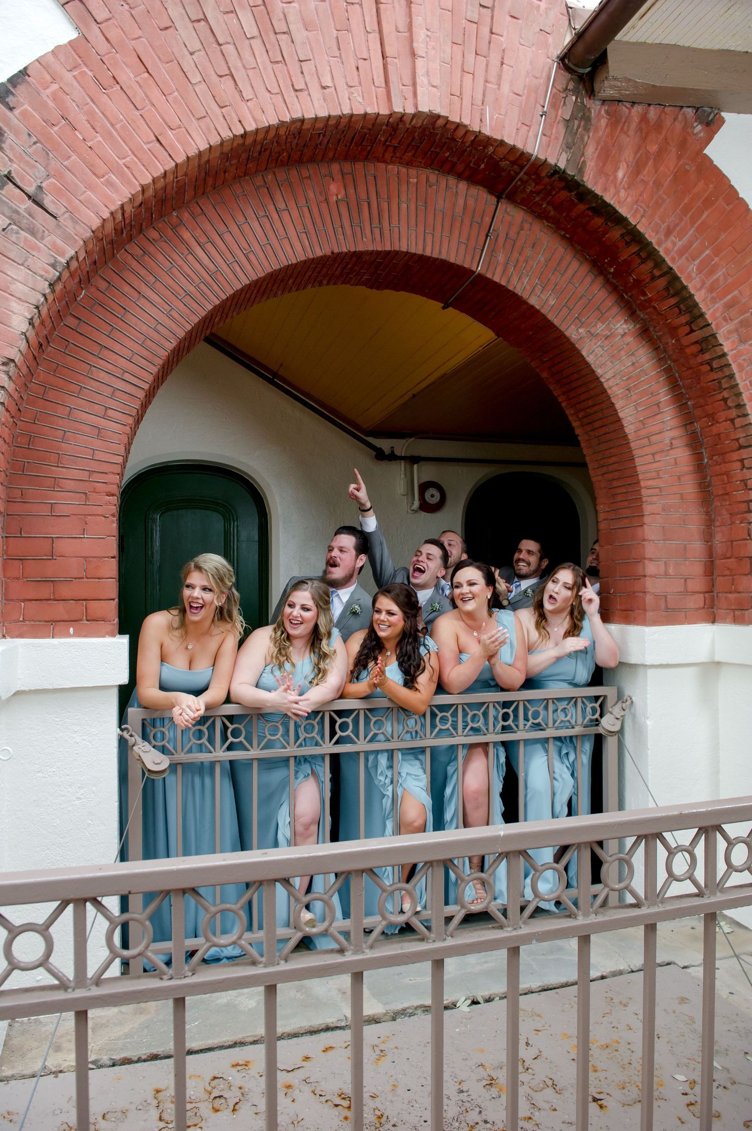 Bridesmaids in blue dresses lean on a balcony railing under a brick arch.