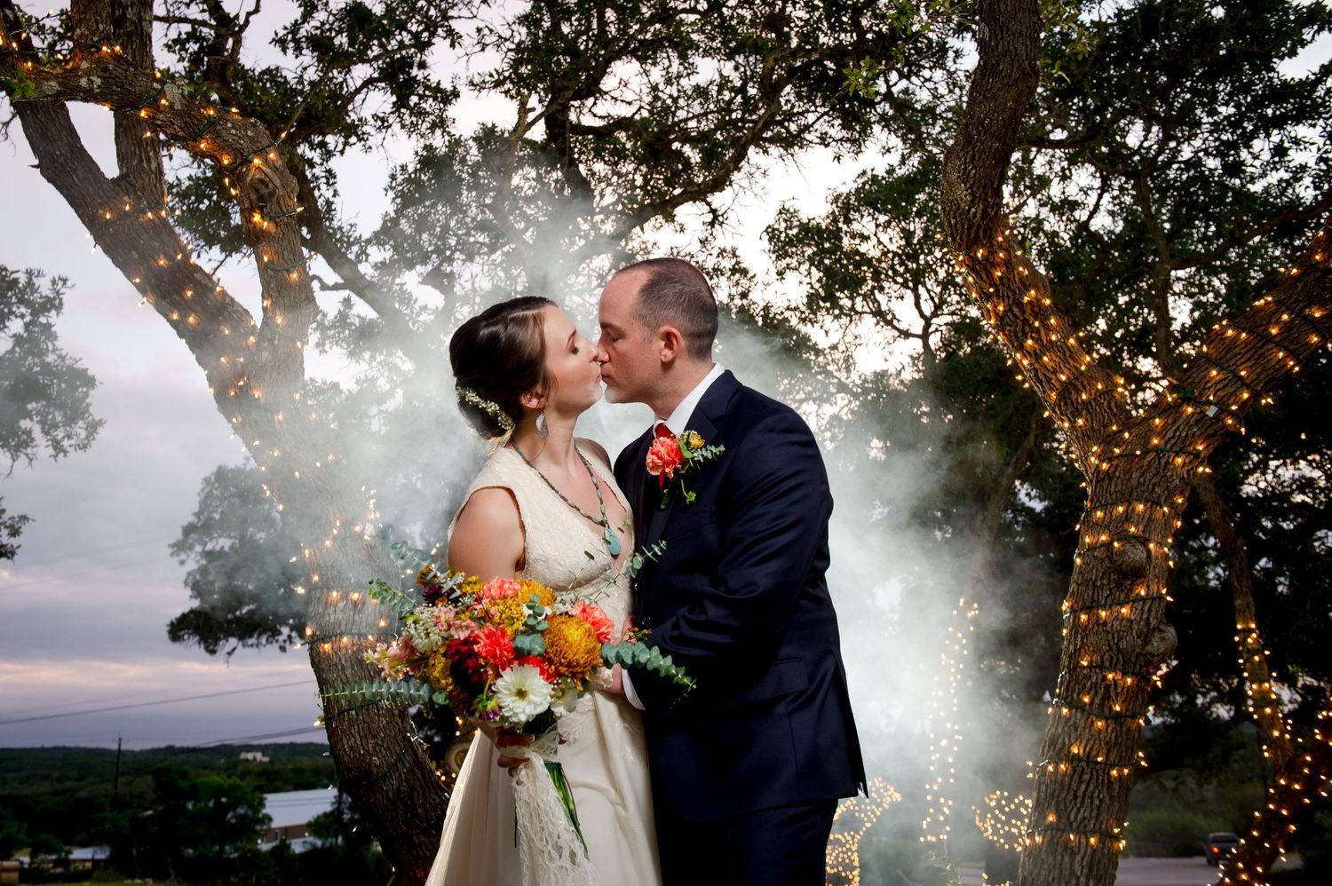 Wedding couple shares intimate moments under twinkling lights and tree branches at dusk.