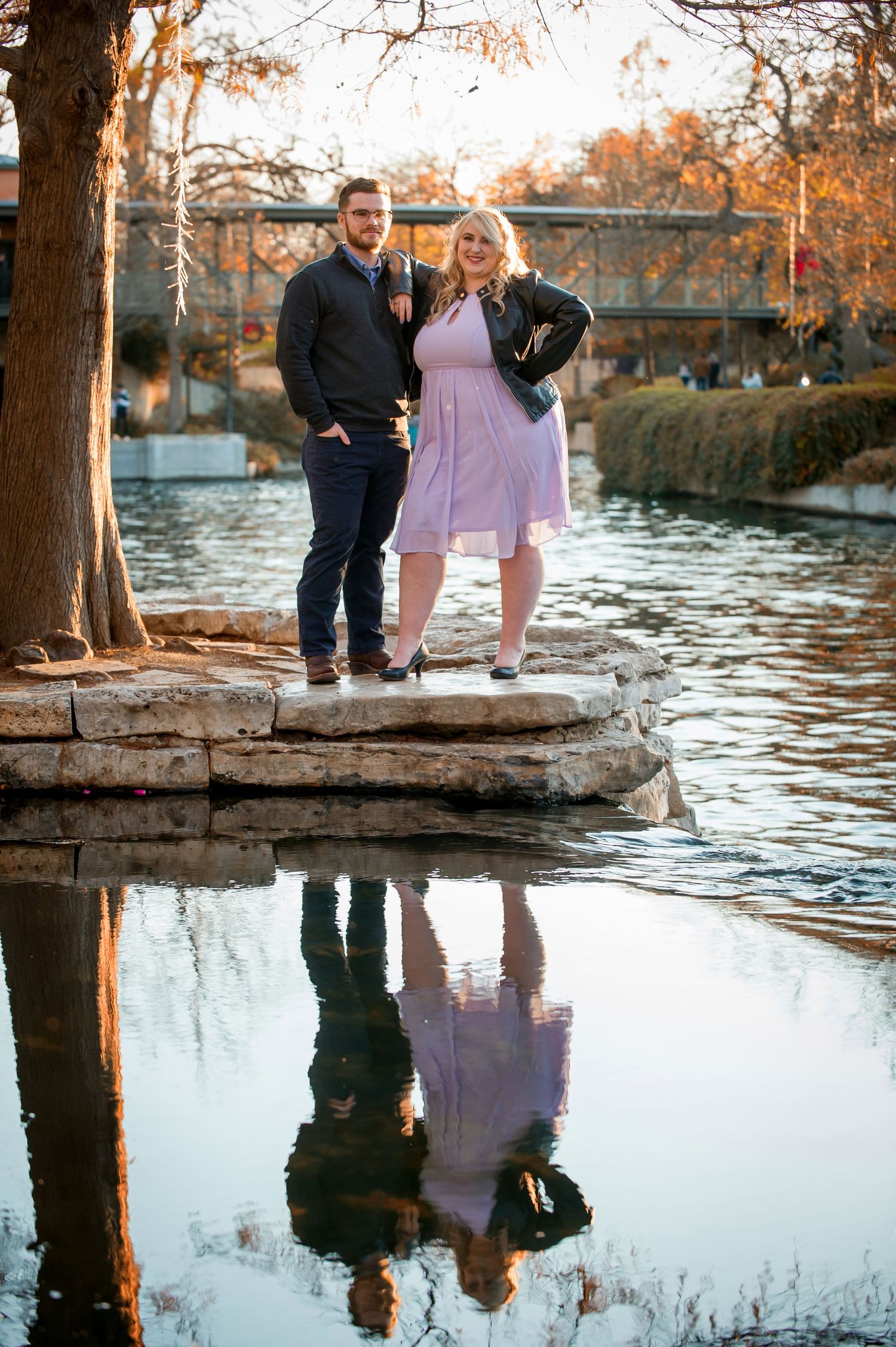 Couple standing on stone ledge by water with autumn trees and building reflection.