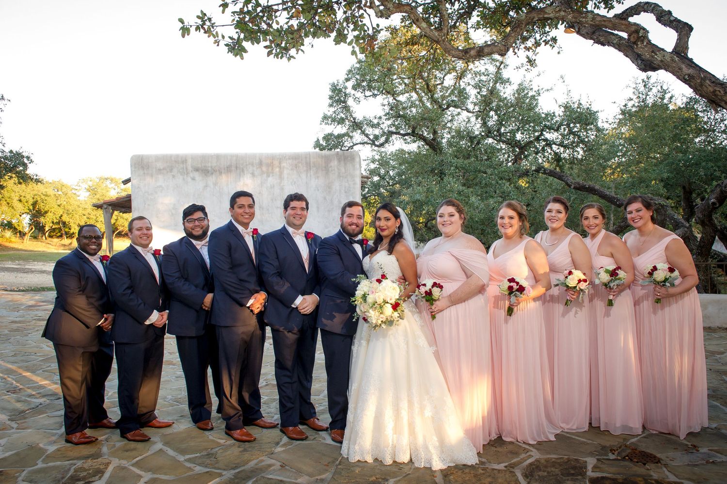 Wedding party in navy and pink attire poses outdoors under large oak trees at sunset.