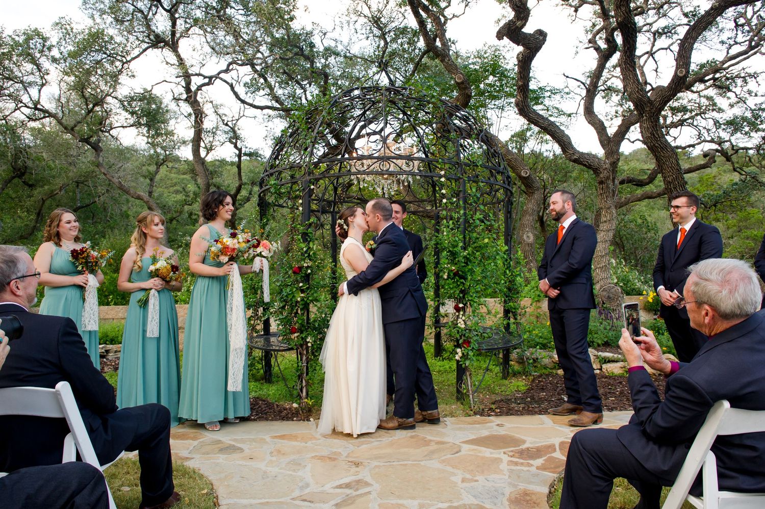 Wedding ceremony guests gathered outdoors under trees at a rustic venue with white chairs.