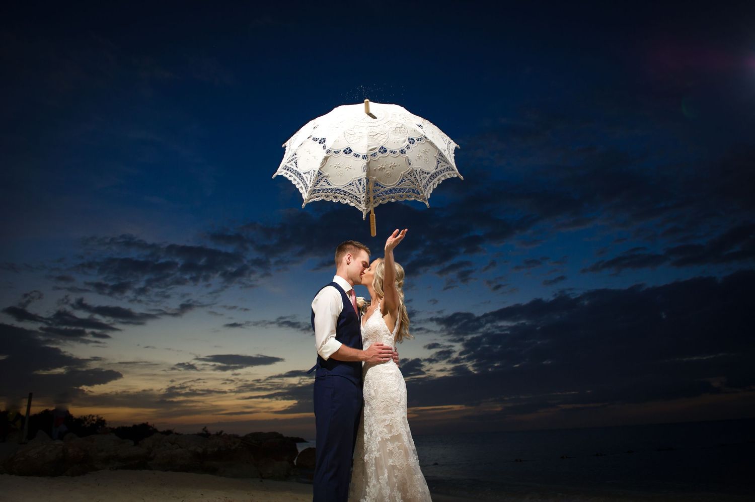 Romantic sunset wedding portraits with white parasol creating silhouettes against dramatic sky.