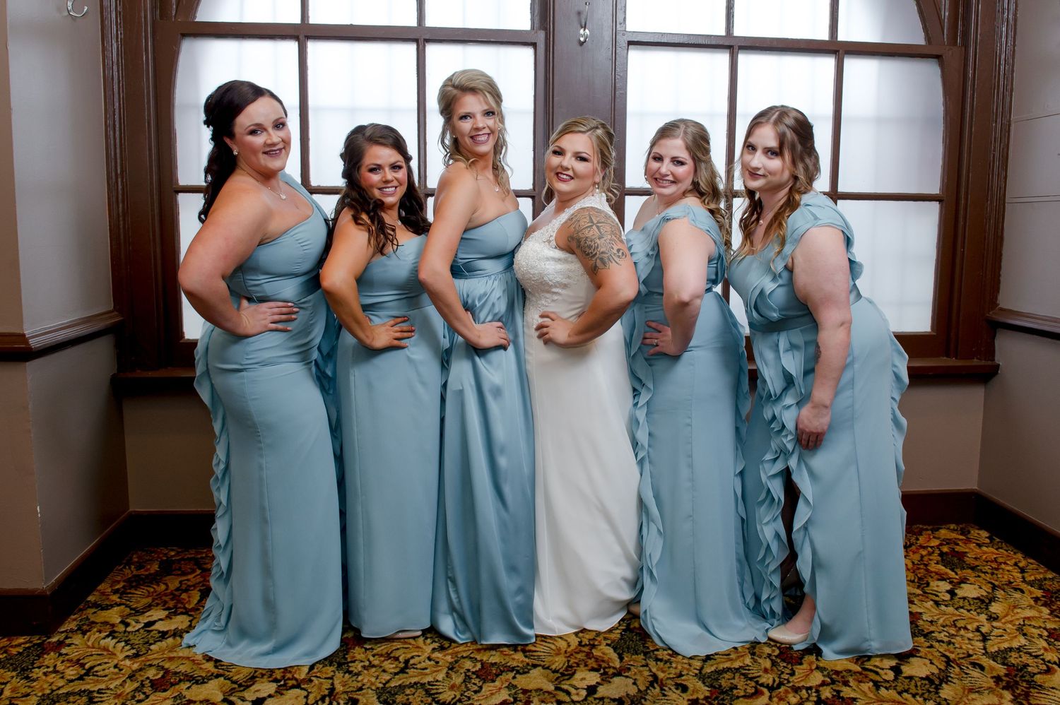 A wedding party in light blue dresses poses together in front of a window.