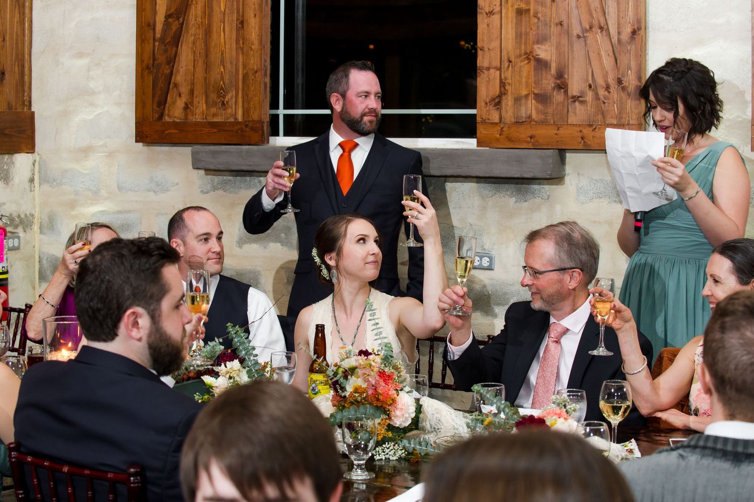 Wedding guests enjoy a lively reception dinner at a rustic venue with exposed wooden beams and warm lighting.