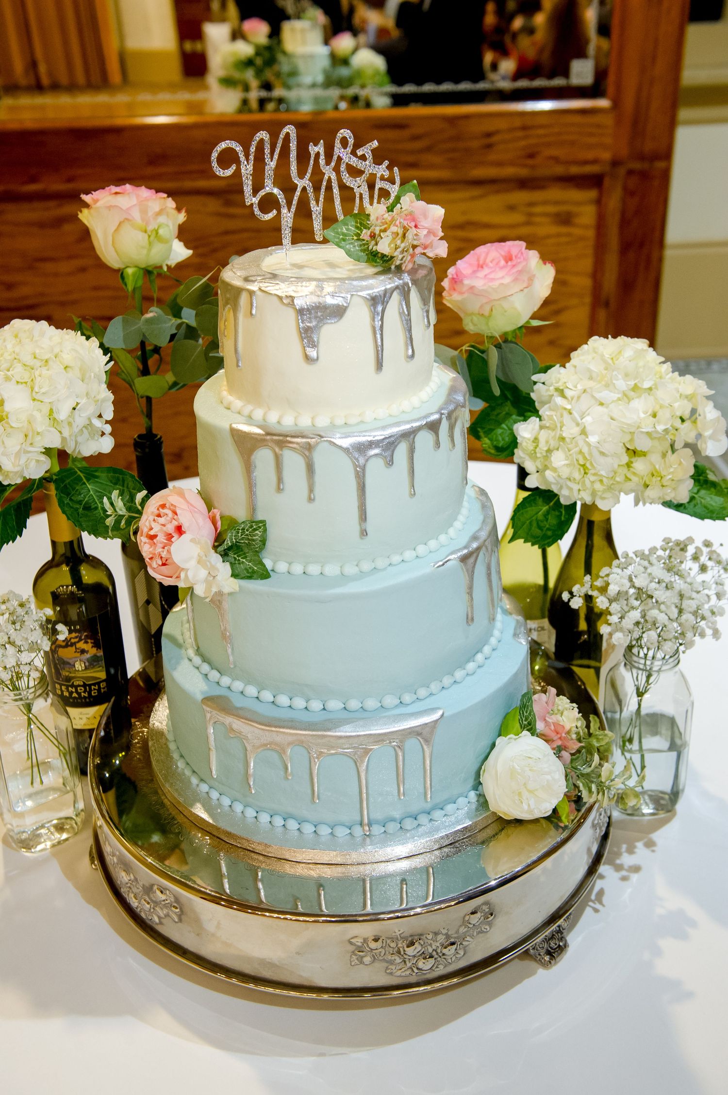 Four-tier light blue wedding cake with silver drip decoration and pink floral accents.