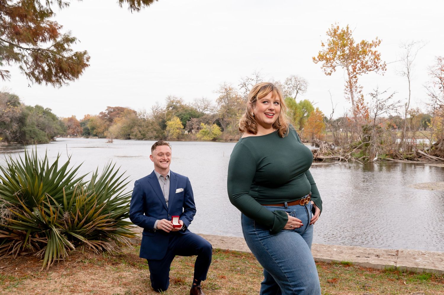 A couple shares a playful moment by a serene lake surrounded by autumn trees.