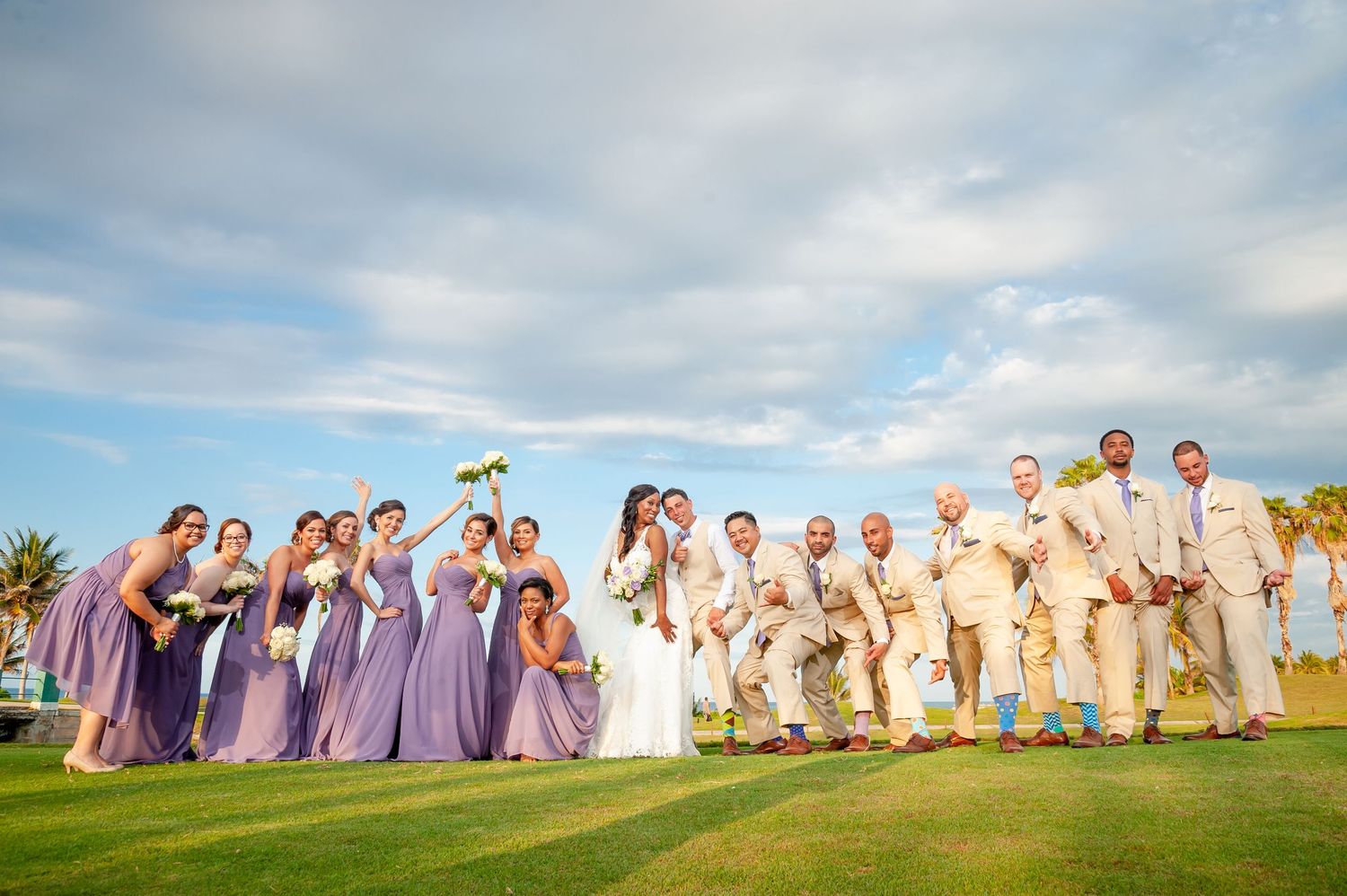 Wedding party in purple dresses and tan suits poses on a scenic golf course under blue skies.