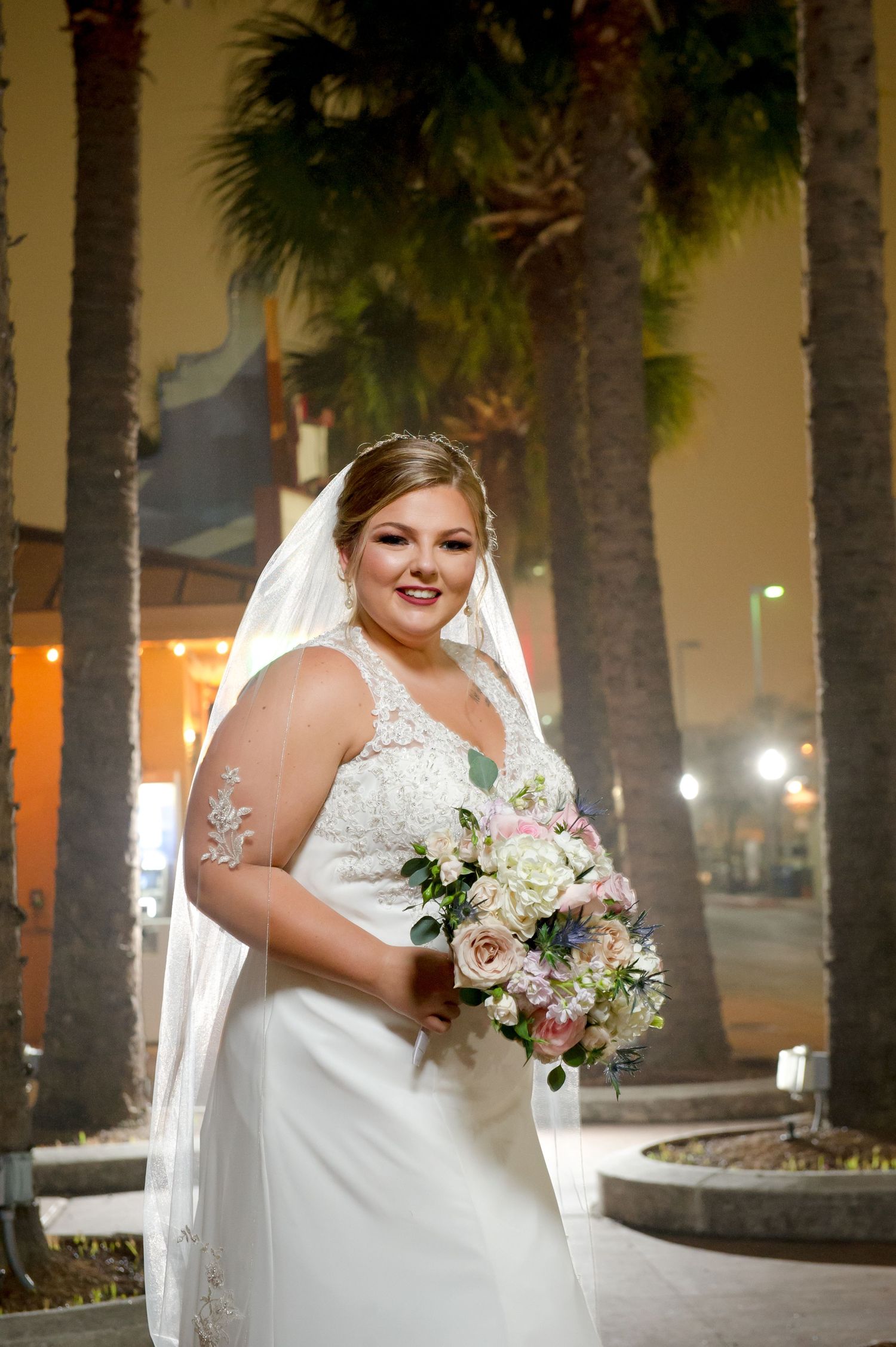 A bride in a white lace dress holds a beautiful bouquet of pink and white flowers against palm trees.