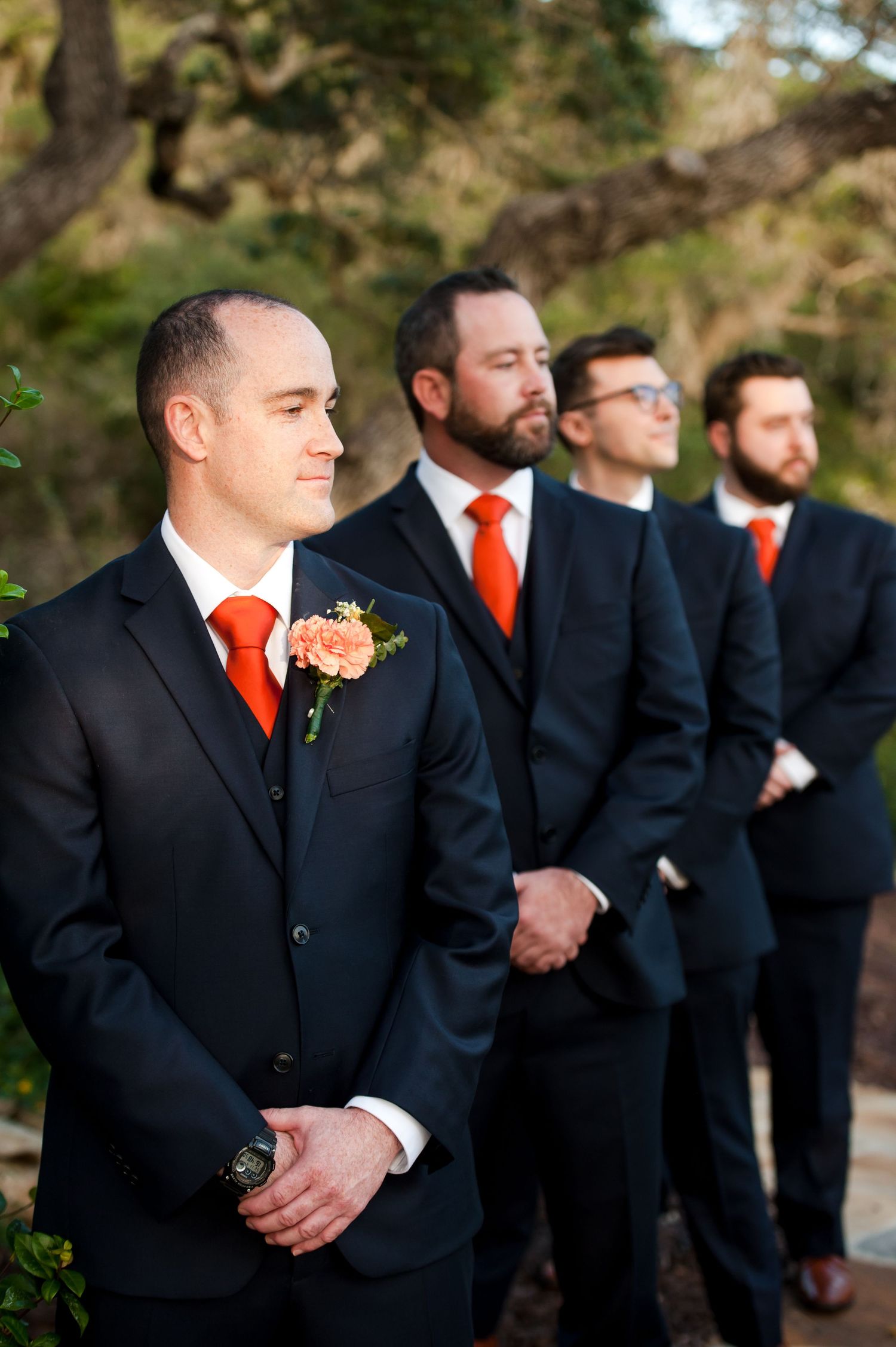 Groomsmen in black suits with bright orange ties and boutonnieres stand in a line outdoors.