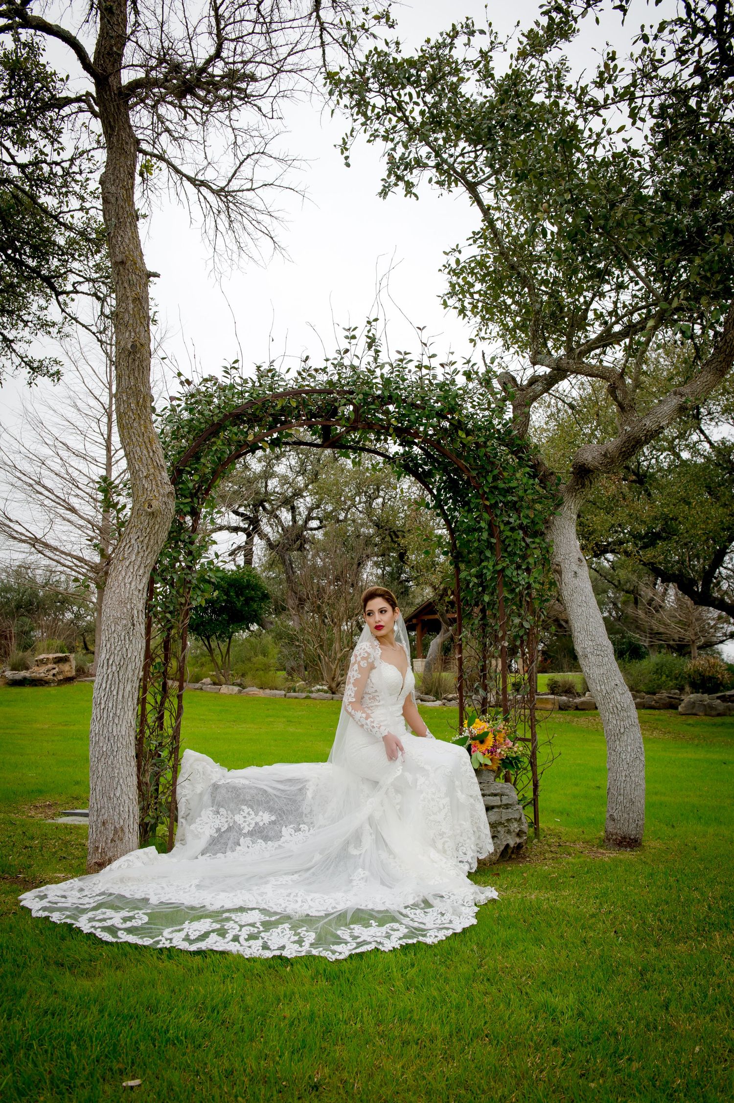 A figure in a flowing white wedding gown and veil poses gracefully among tall trees in a lush green outdoor setting.