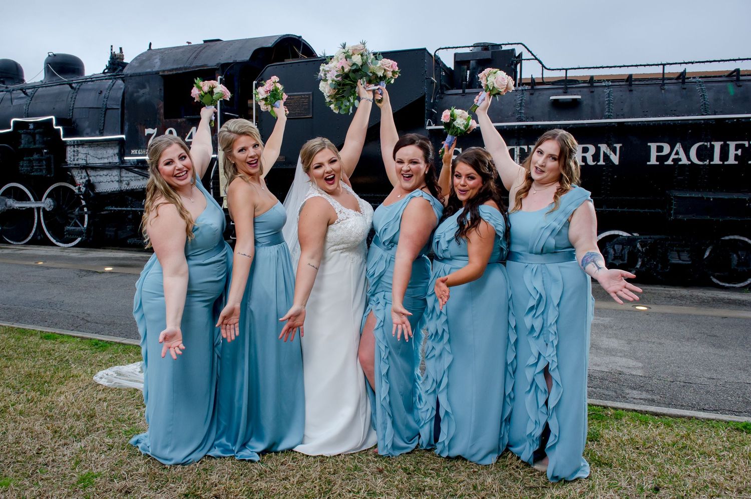 Bridal party in blue dresses celebrates joyfully by vintage locomotive.