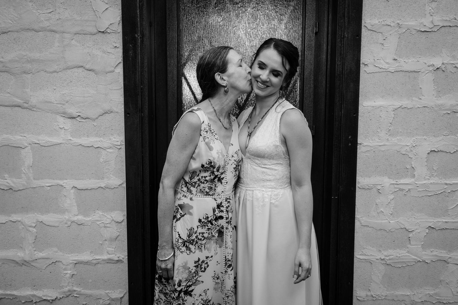 Two people in formal attire share a candid moment laughing together against a white brick wall.