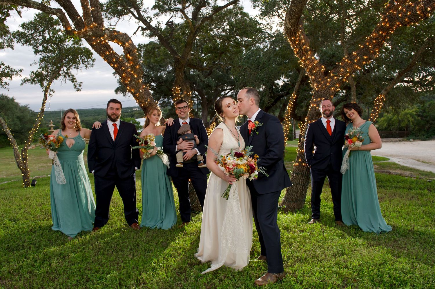Wedding party in turquoise dresses and dark suits posed under trees with string lights.