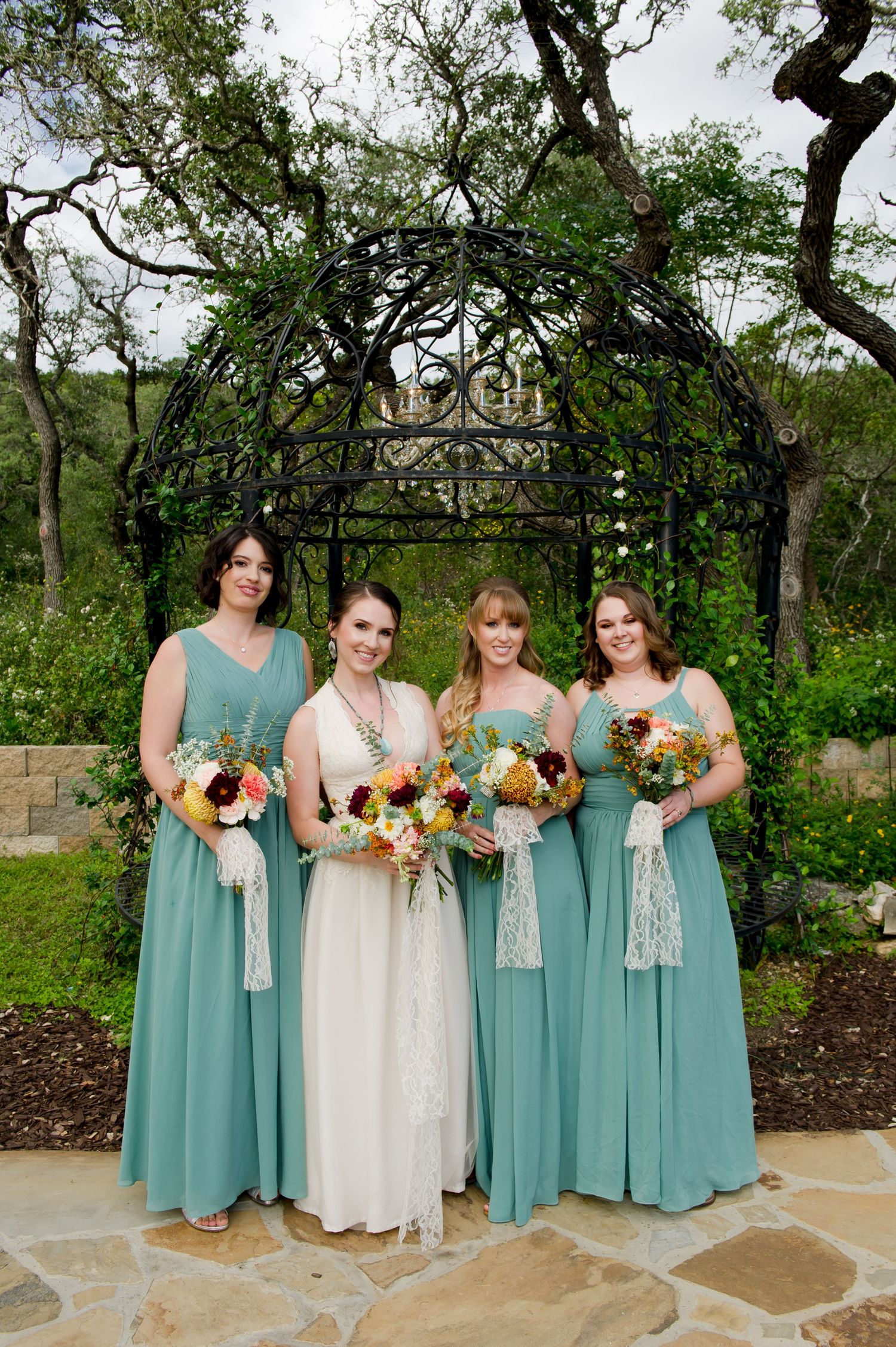 A wedding party in seafoam green dresses holding colorful bouquets poses under a garden arbor on stone patio.