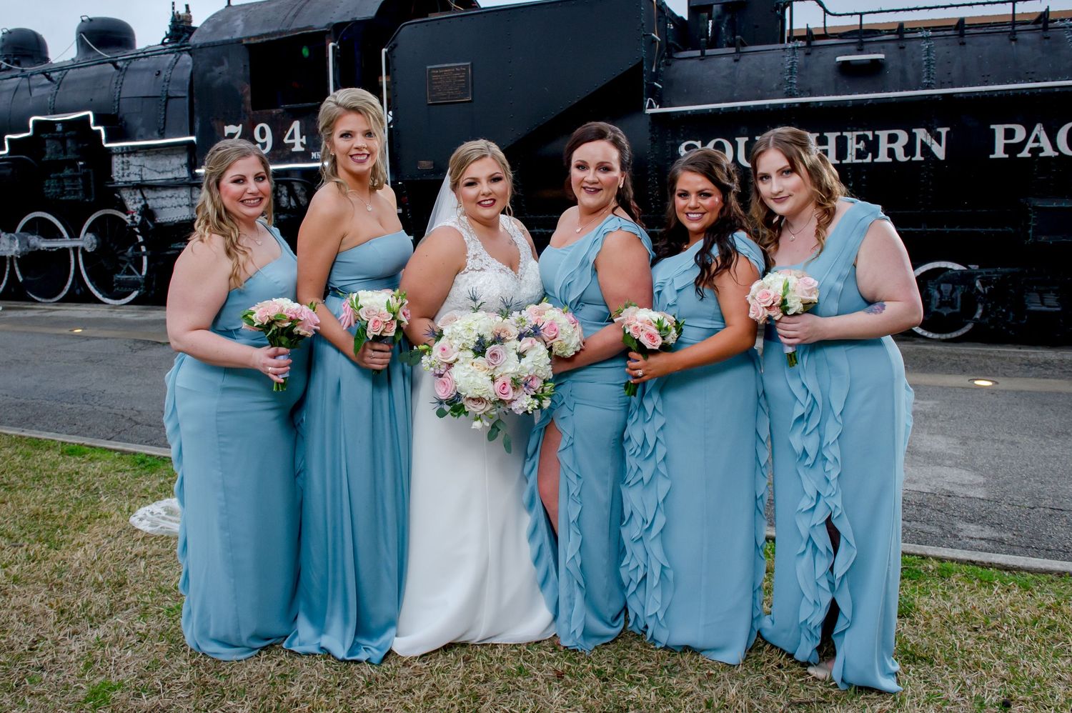 Wedding party in light blue dresses and white gown pose in front of vintage Southern Pacific train.