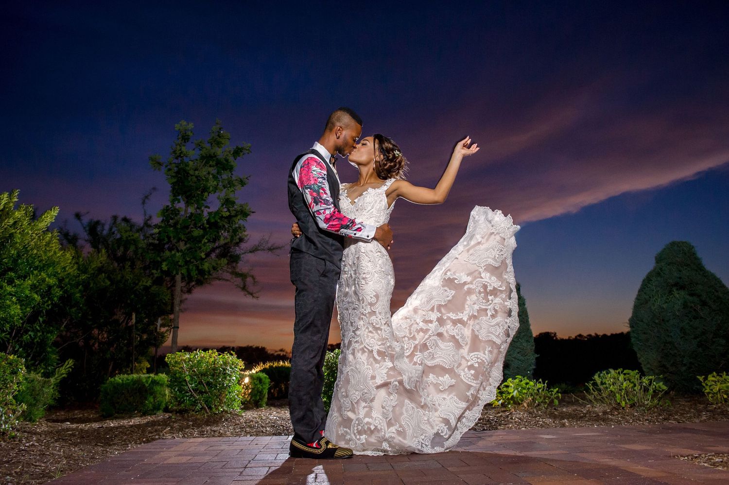 A couple shares a romantic moment at sunset, with the bride's lace dress flowing dramatically against the colorful sky.