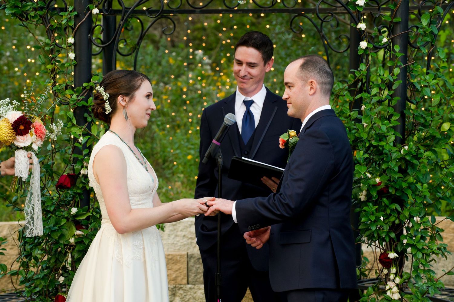 Wedding ceremony taking place under a garden archway decorated with twinkling lights and greenery.