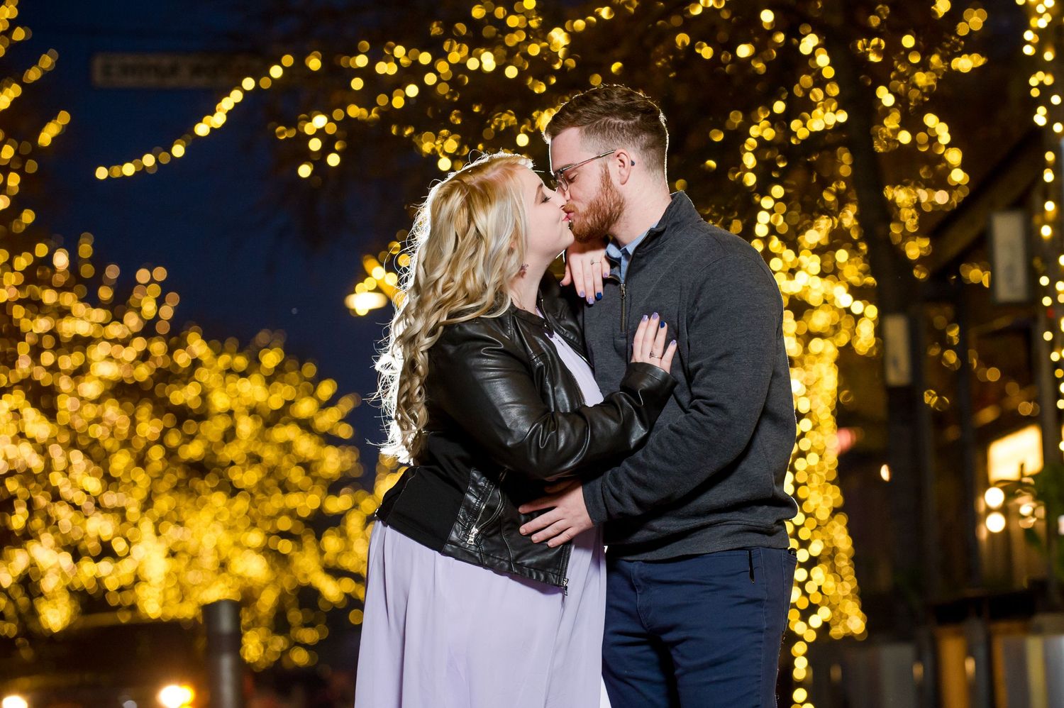 Romantic couple shares a kiss under twinkling golden lights at twilight during their wedding celebration.