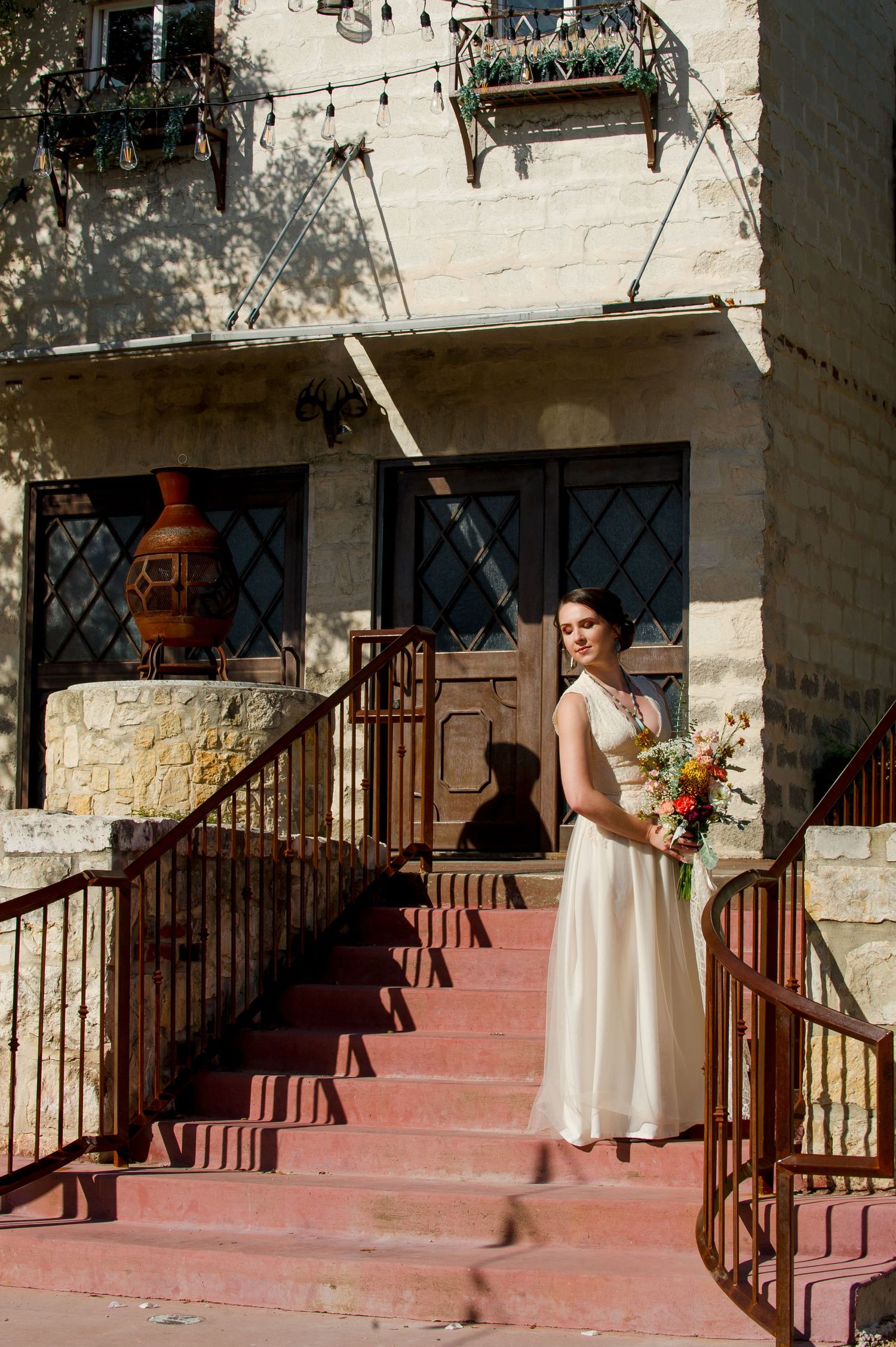 A person in a white dress stands on stone steps with a rustic wooden railing and red floor against a light stone wall.