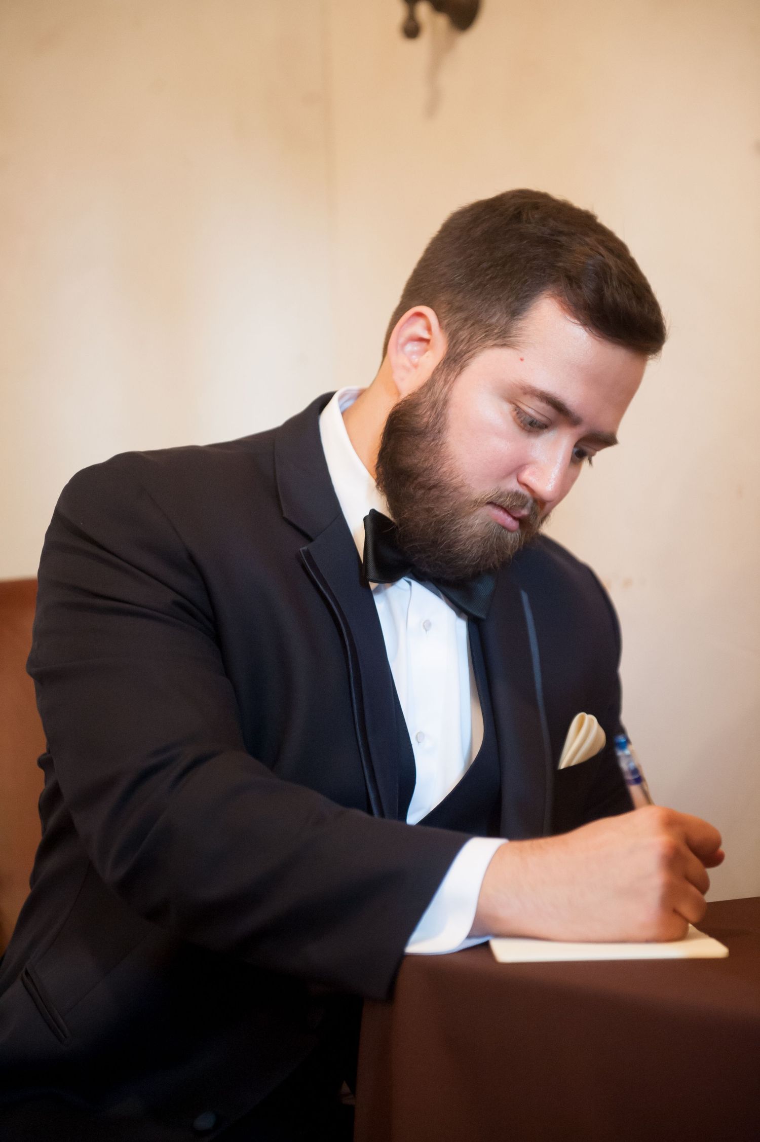 Groom in tuxedo writing thoughtfully at wooden desk in vintage setting.
