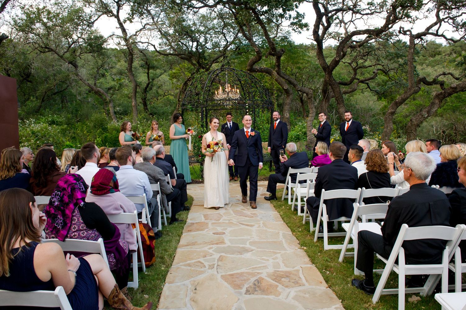 Outdoor wedding ceremony setup with white chairs on stone patio surrounded by trees.