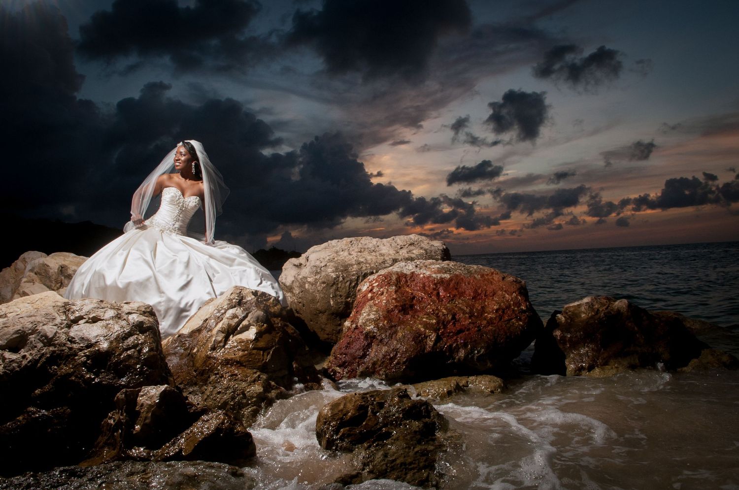 Dramatic bridal portrait on coastal rocks at sunset with crashing waves.