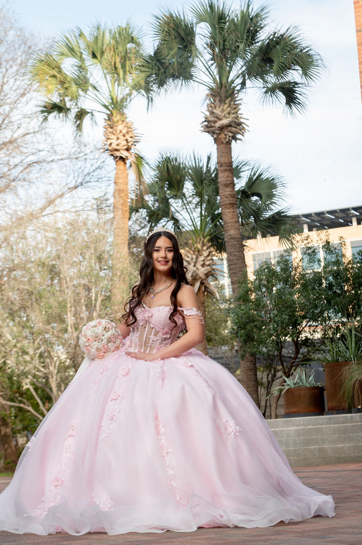 Pink quinceañera dress showcased in outdoor setting with palm trees.