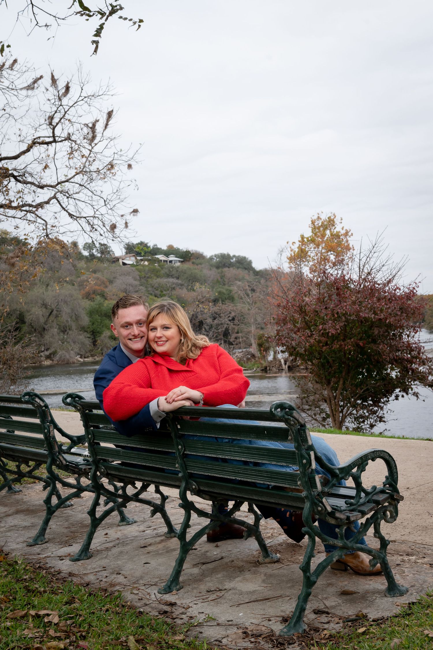 A couple relaxes on a green metal park bench with a lake view.