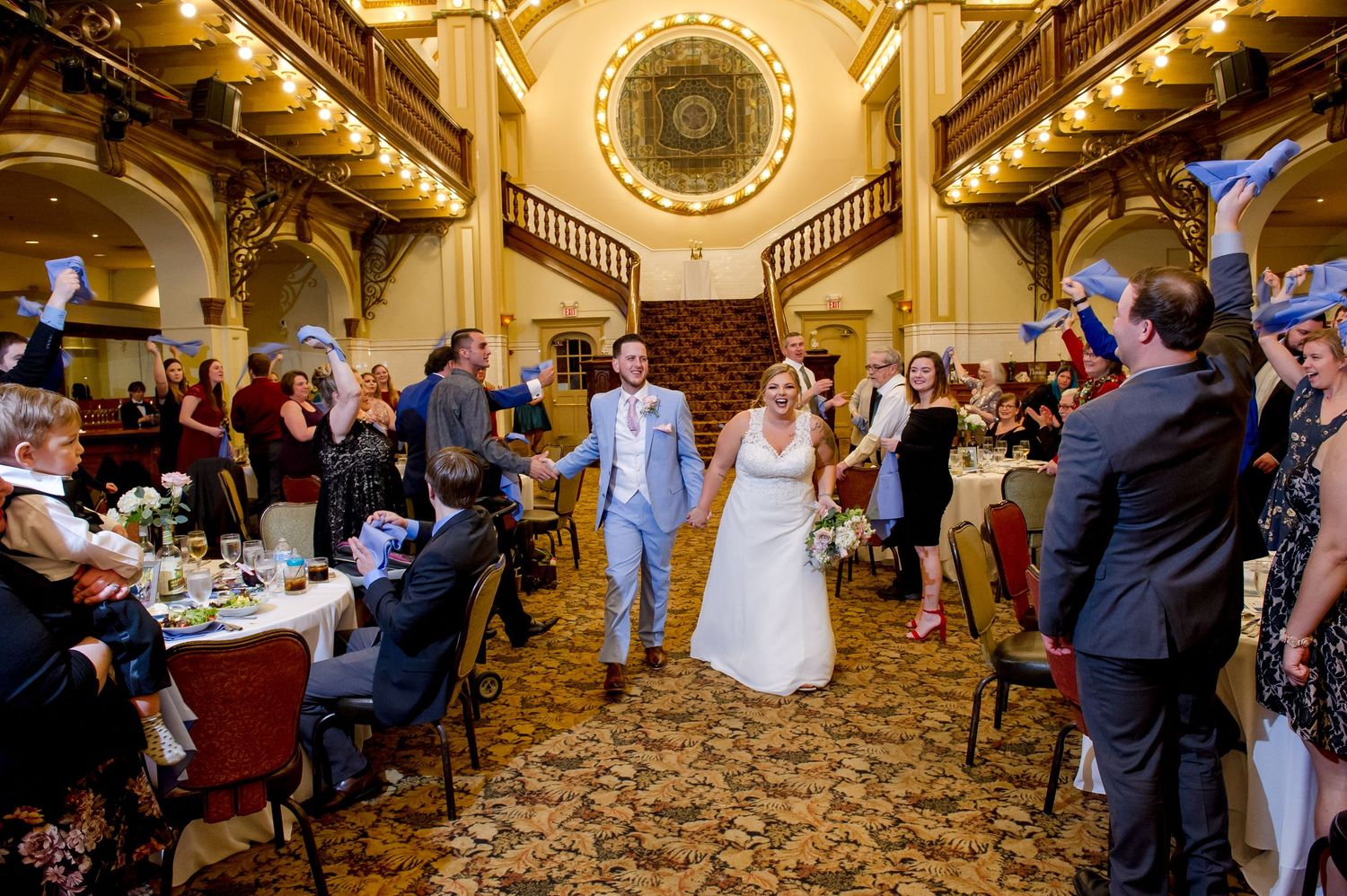 Newlyweds exit ornate ballroom while guests celebrate on both sides.
