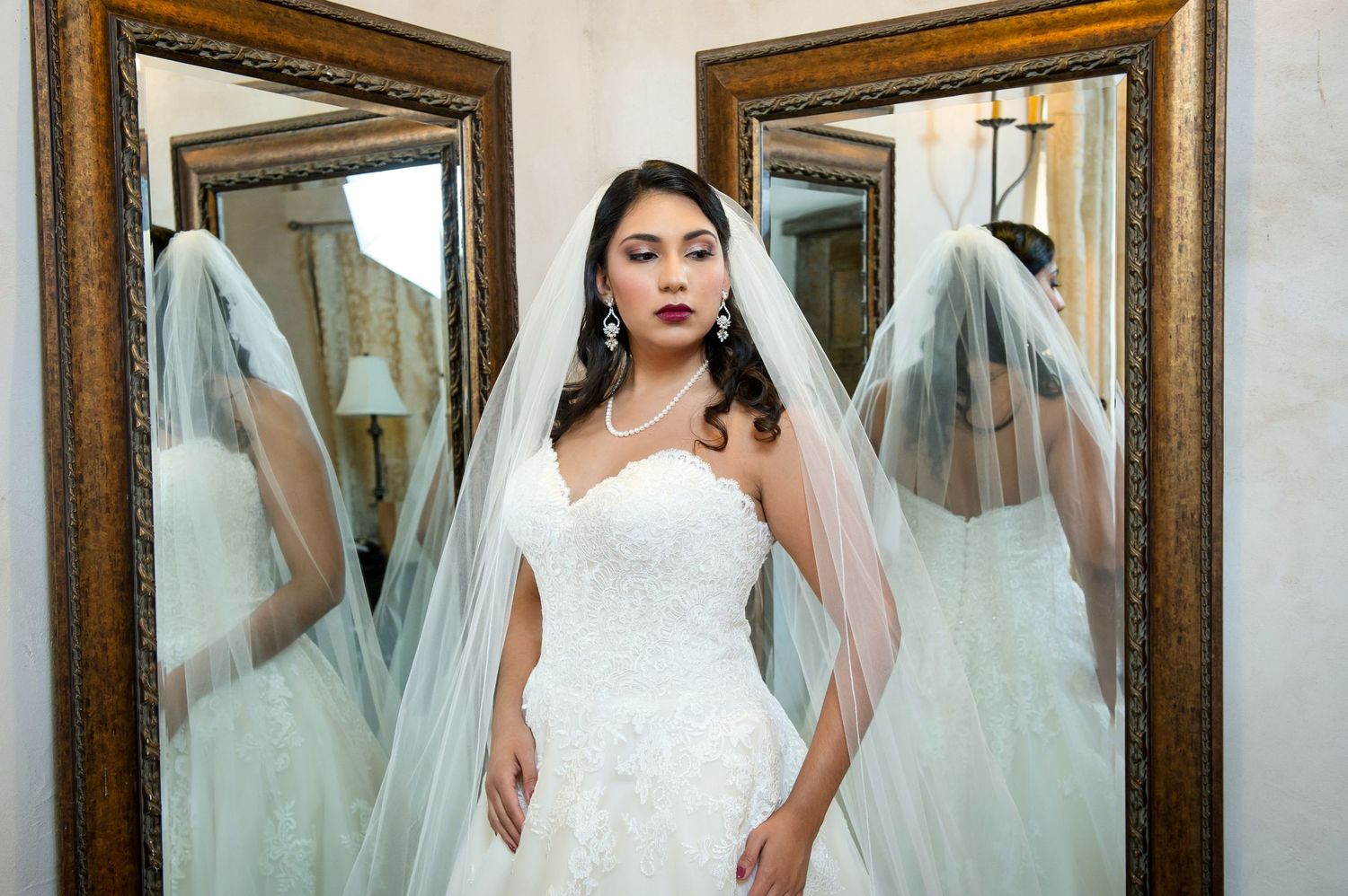 Dramatic mirror reflection of bride in fitted lace gown with flowing veil in ornate gold frames.