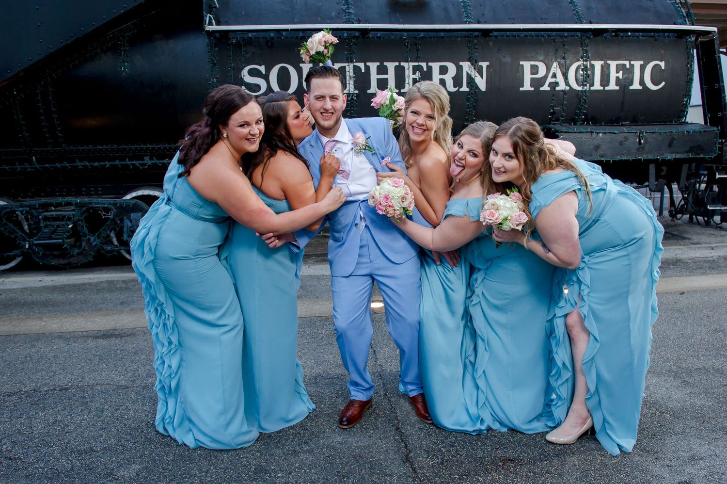 Wedding party in light blue dresses poses playfully in front of a vintage Southern Pacific train.