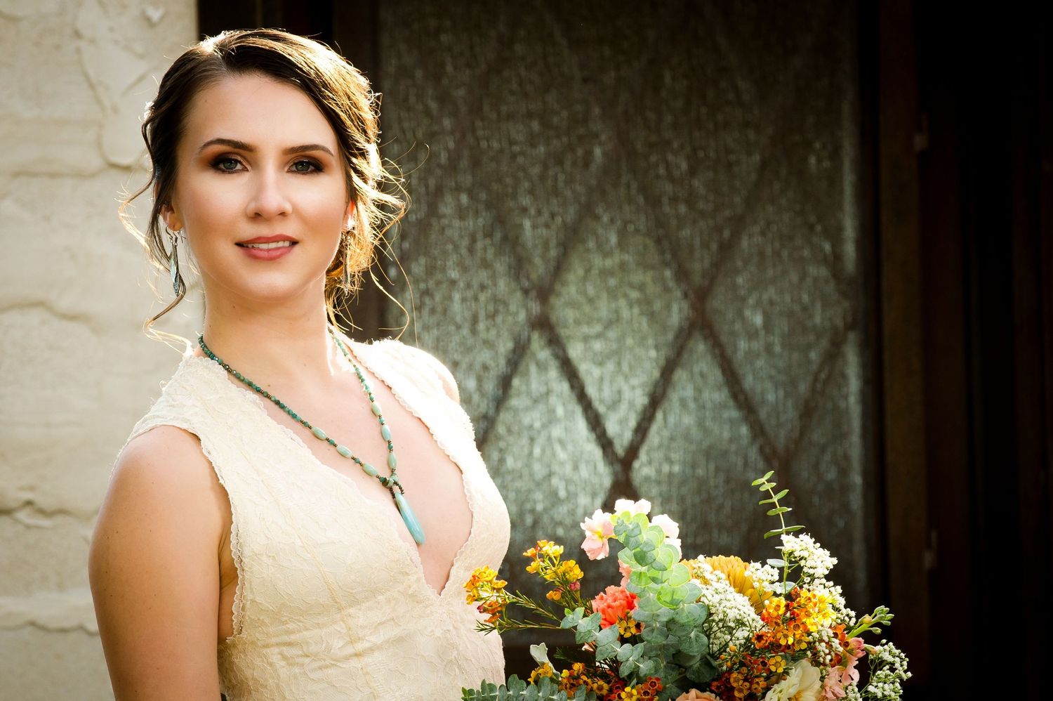 A bride in an elegant cream wedding dress poses with a colorful autumn bouquet against a rustic stone wall.