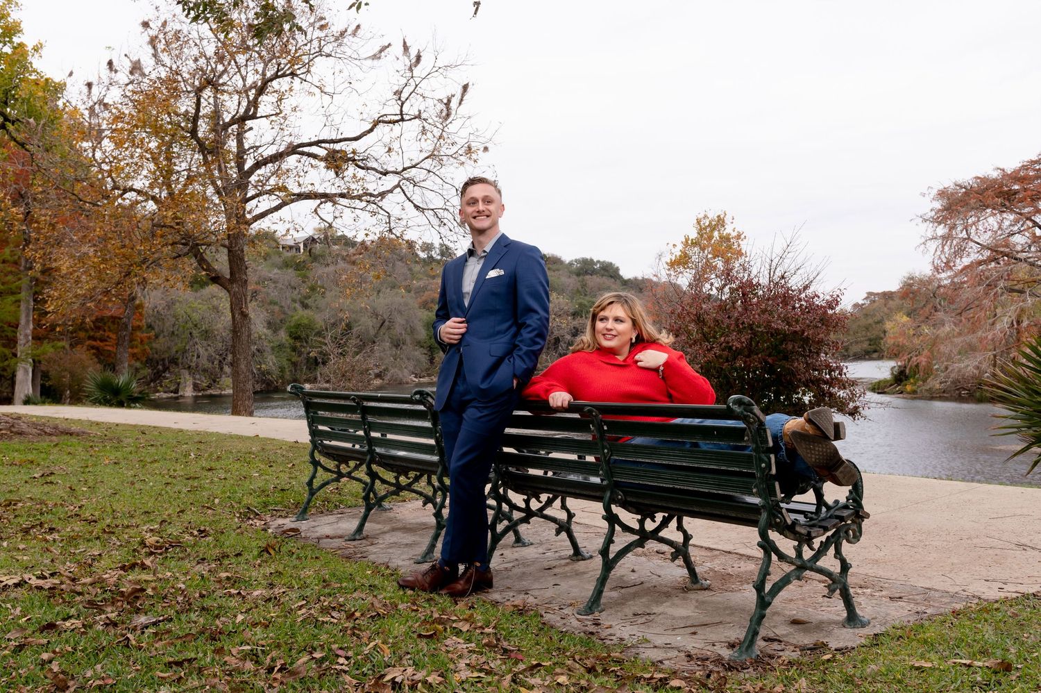 A couple poses playfully on a park bench surrounded by fall foliage.
