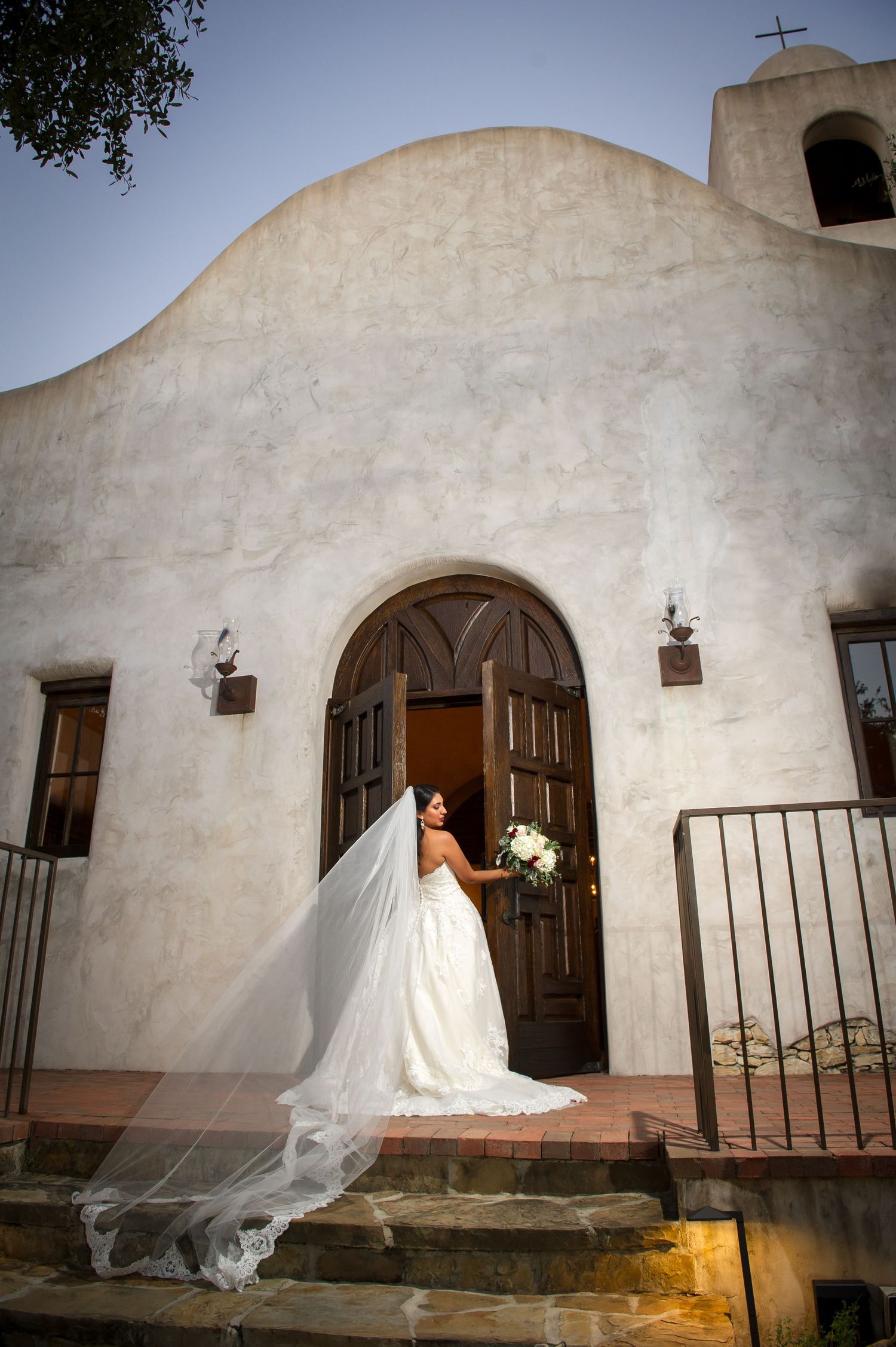 White stucco mission-style church exterior with wooden doors and flowing bridal veil in foreground.