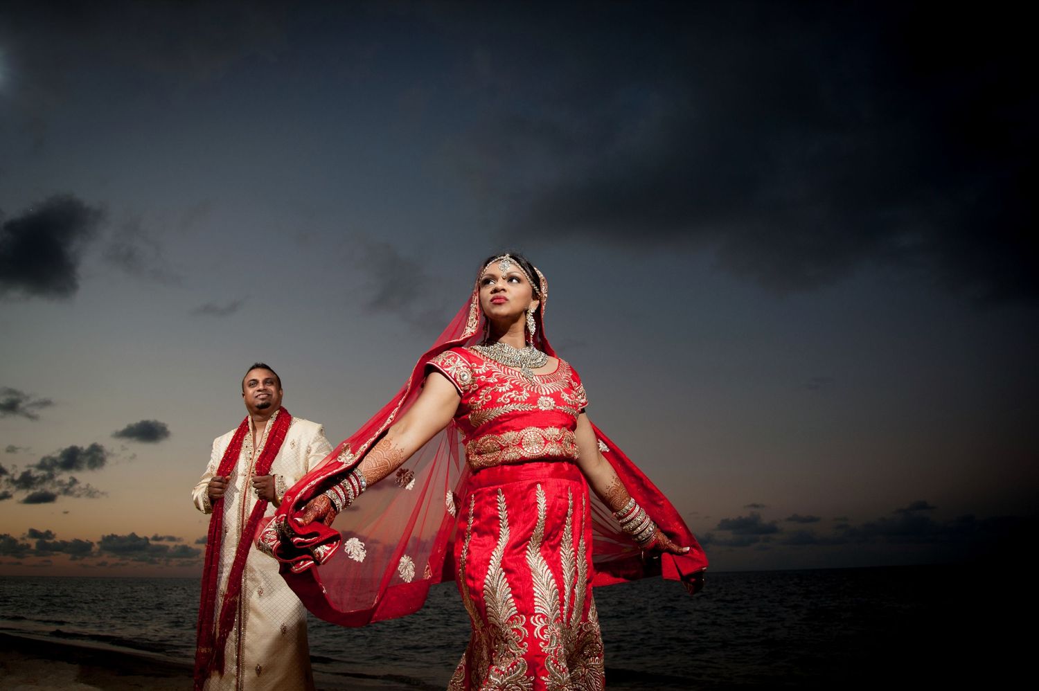 Traditional Indian wedding couple in red attire poses dramatically at sunset by the ocean.