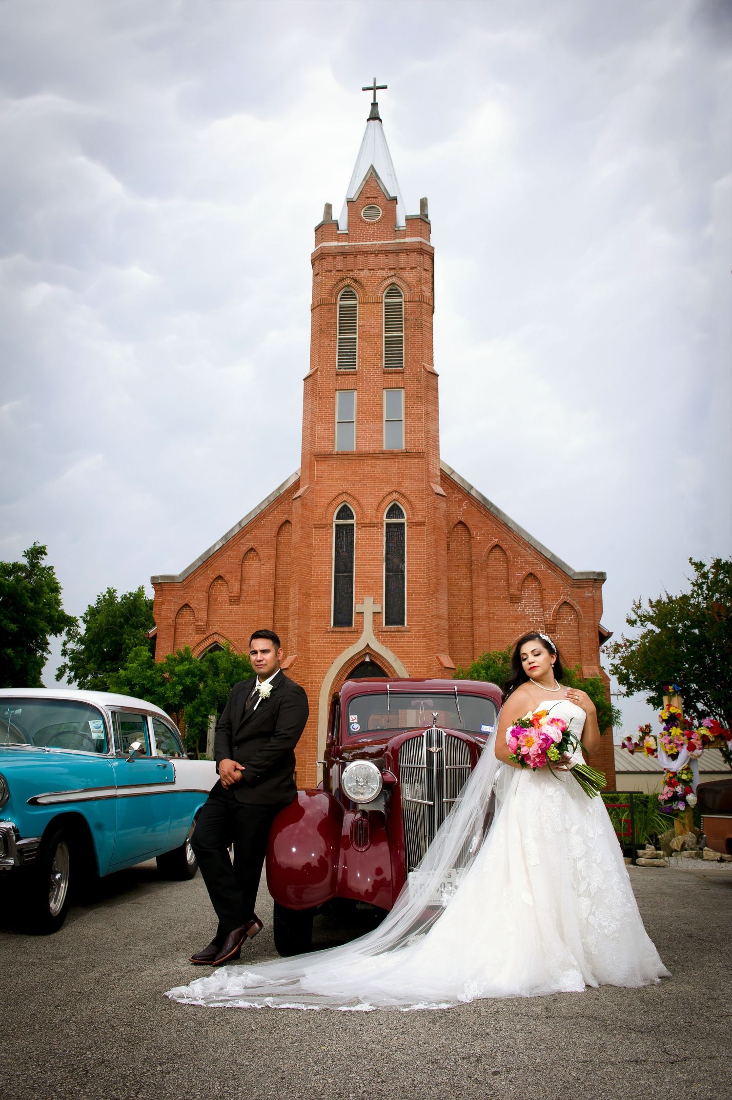 Wedding party gathered outside historic red brick church with vintage cars in background.