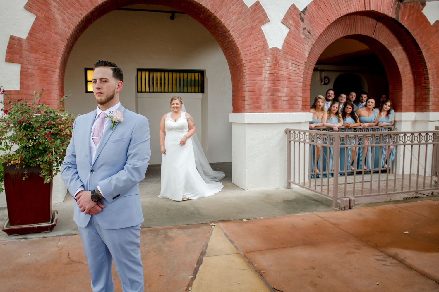A wedding party poses at a brick archway with bridesmaids watching from a balcony.